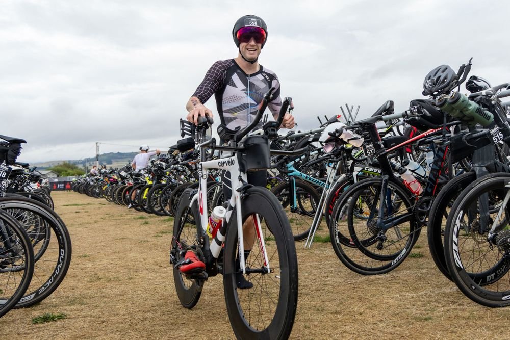 A person in cycling gear and helmet smiling while holding a black and white bicycle at a bike rack filled with other bicycles.