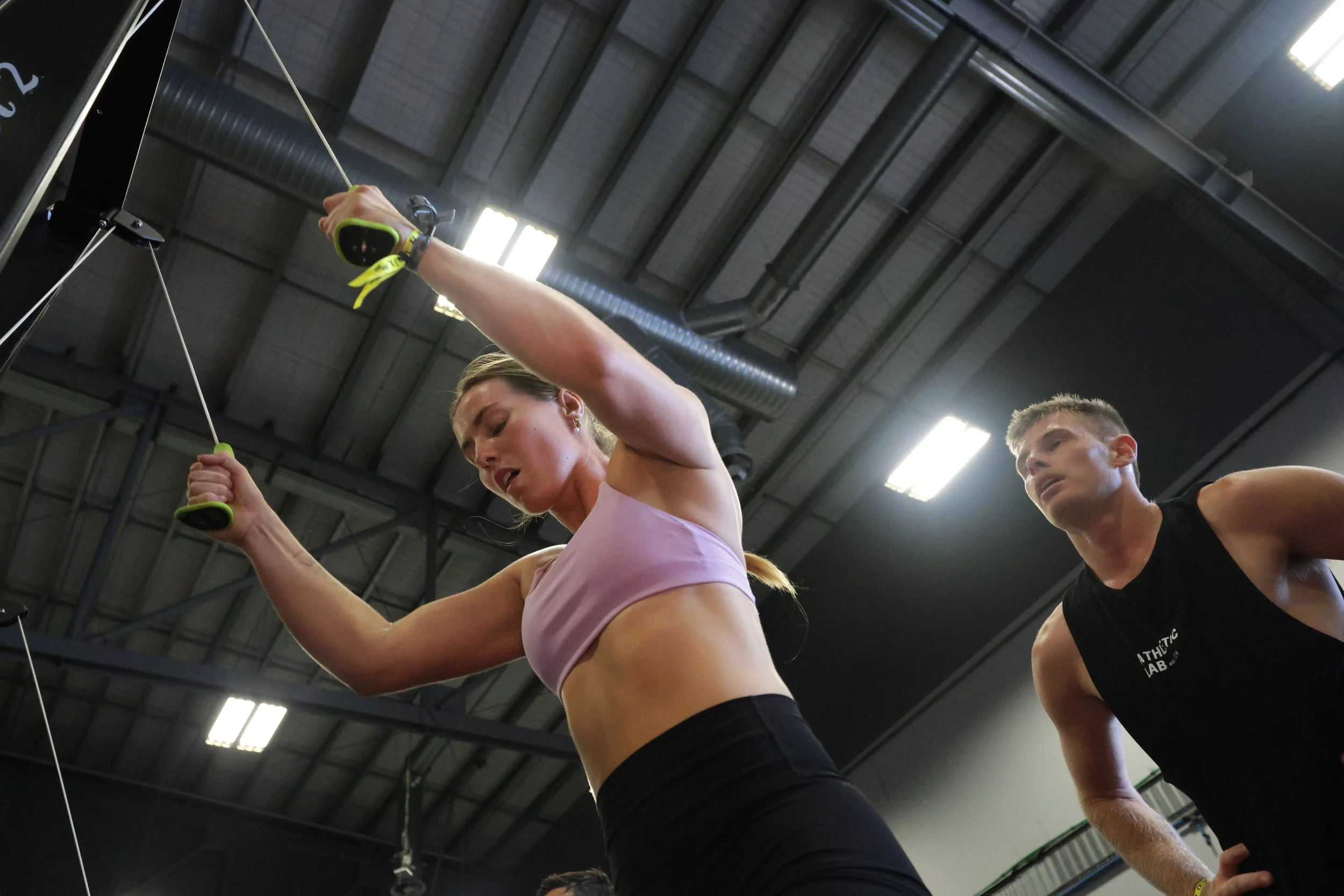 Two people workout at a gym, pulling on resistance bands attached to the ceiling, with gym equipment and ceiling lights visible in the background.