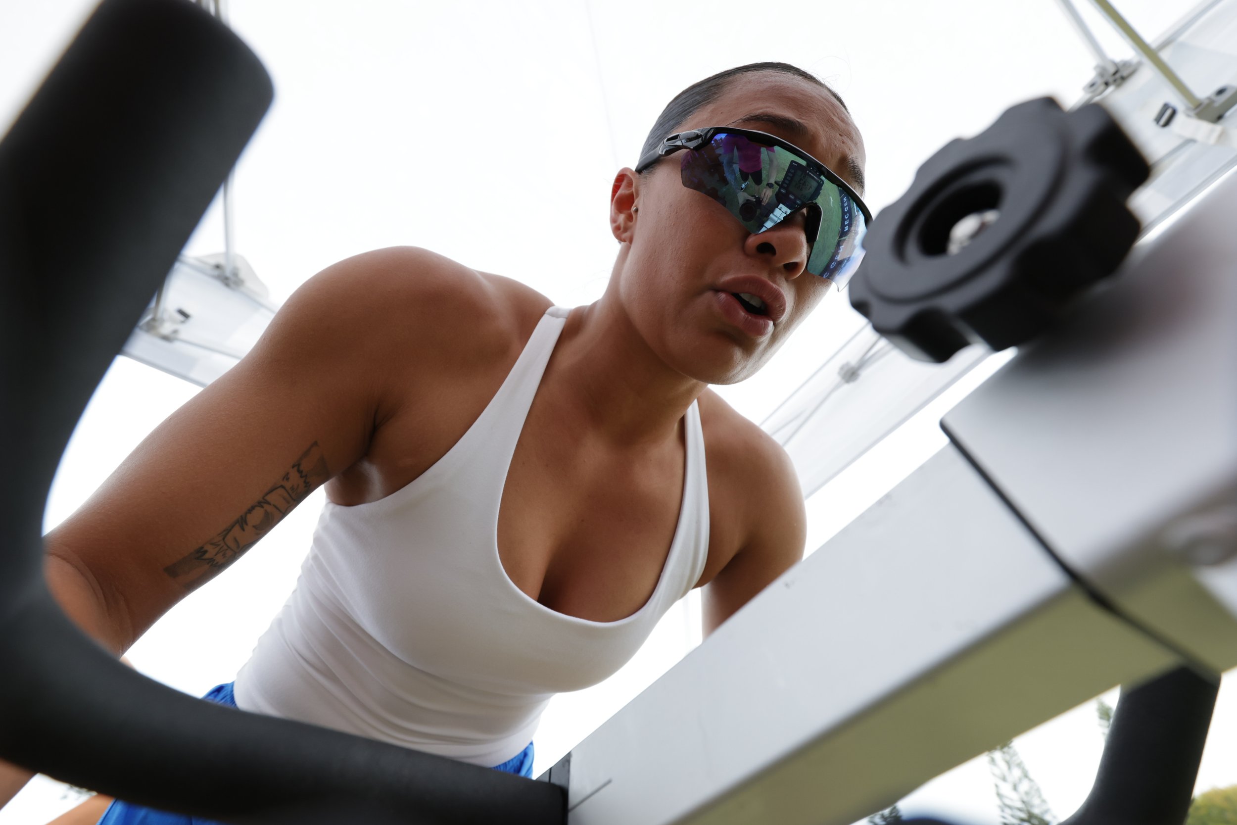 A woman in a white tank top wearing sports sunglasses is looking down at a piece of equipment, possibly a treadmill or exercise machine, seen from a low camera angle.