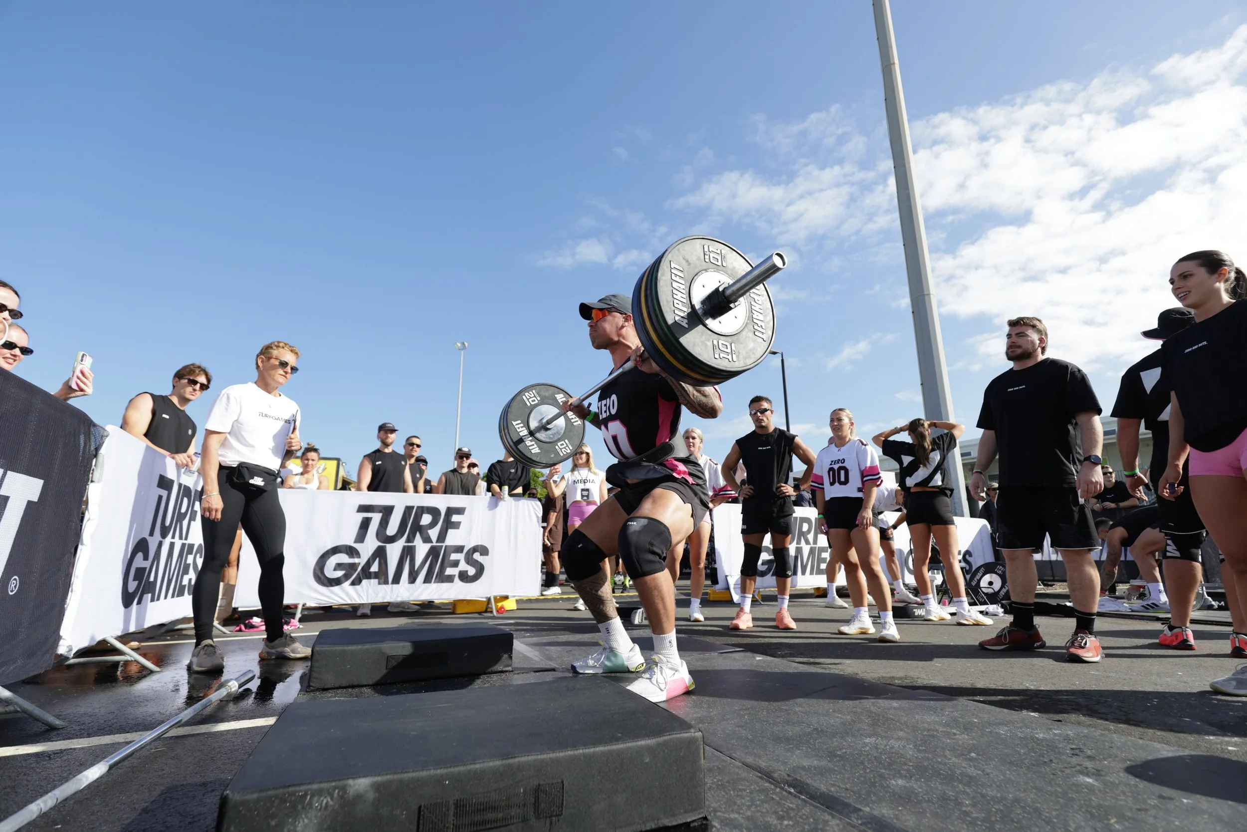A man lifting a barbell with weights during a strength competition, surrounded by spectators on a sunny day.