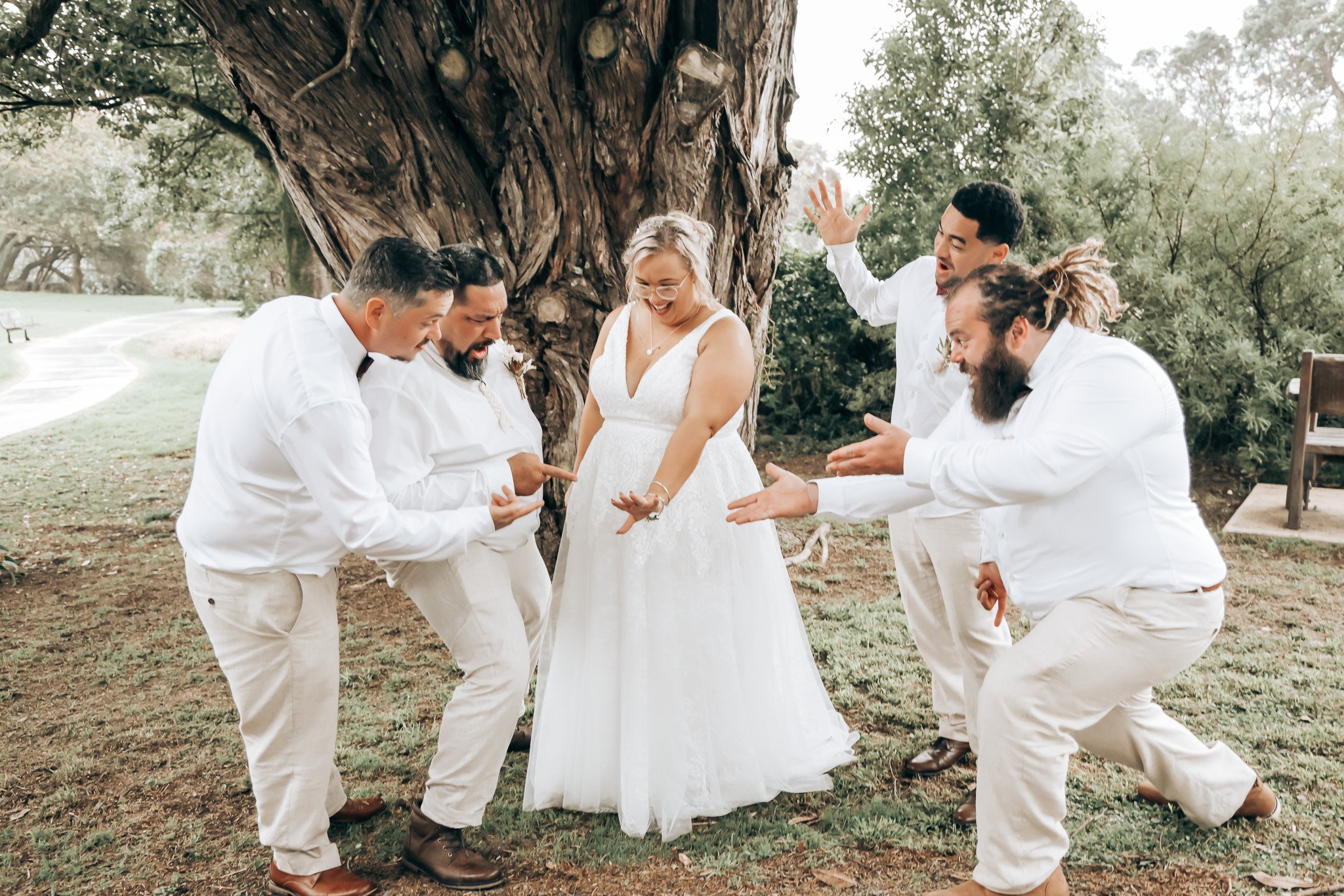 A bride in a white wedding dress and glasses is laughing as she is surrounded by five men dressed in white shirts and beige pants, all engaged in a joyful moment outdoors under a large tree.