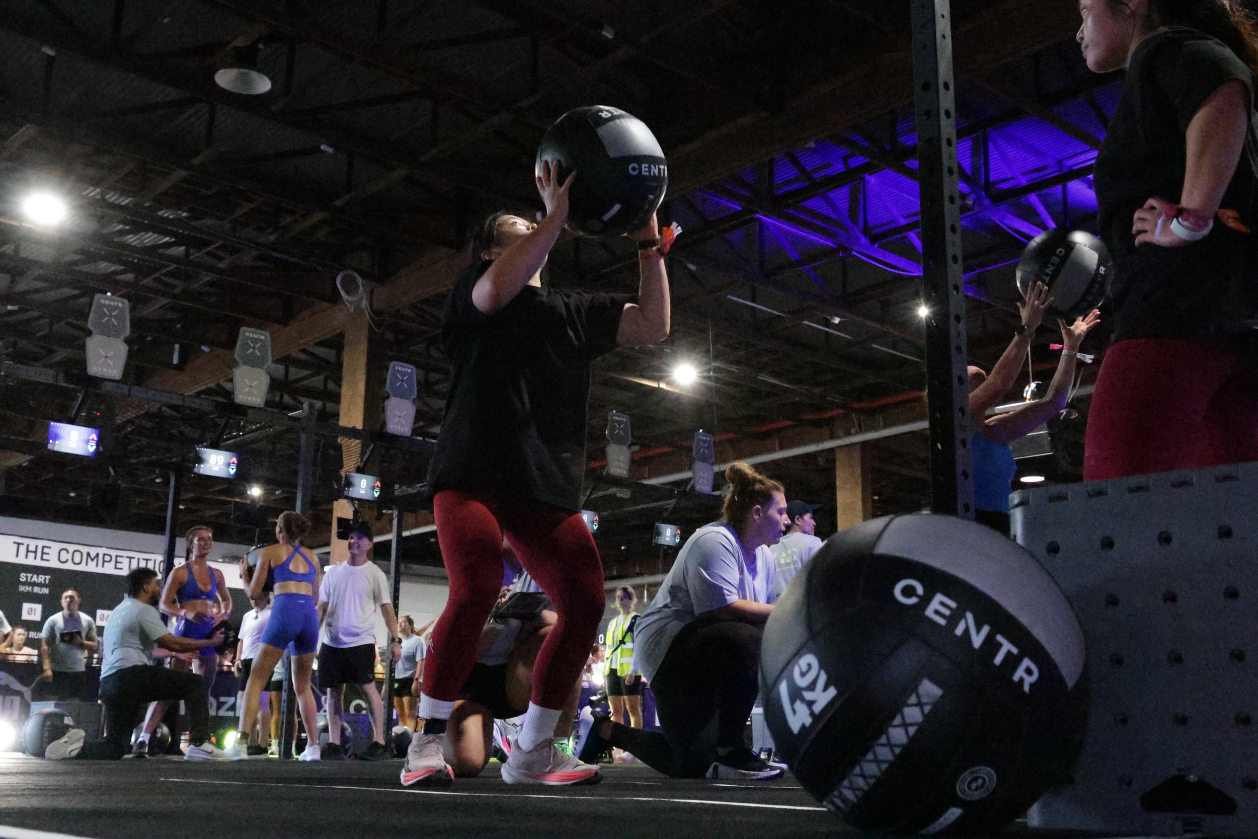 Women during a CrossFit workout in a gym, lifting medicine balls, with other participants and training equipment visible in the background.
