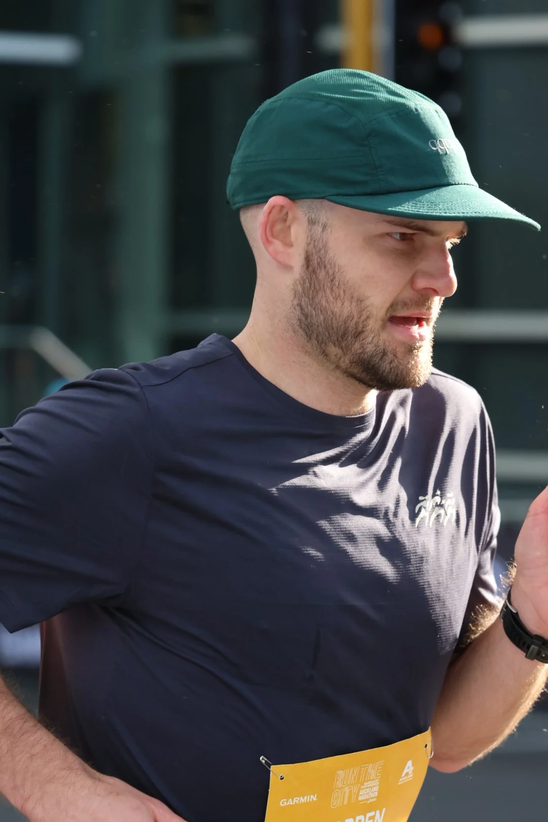 A man running outdoors wearing a green cap, a navy blue athletic shirt, and a yellow race bib.