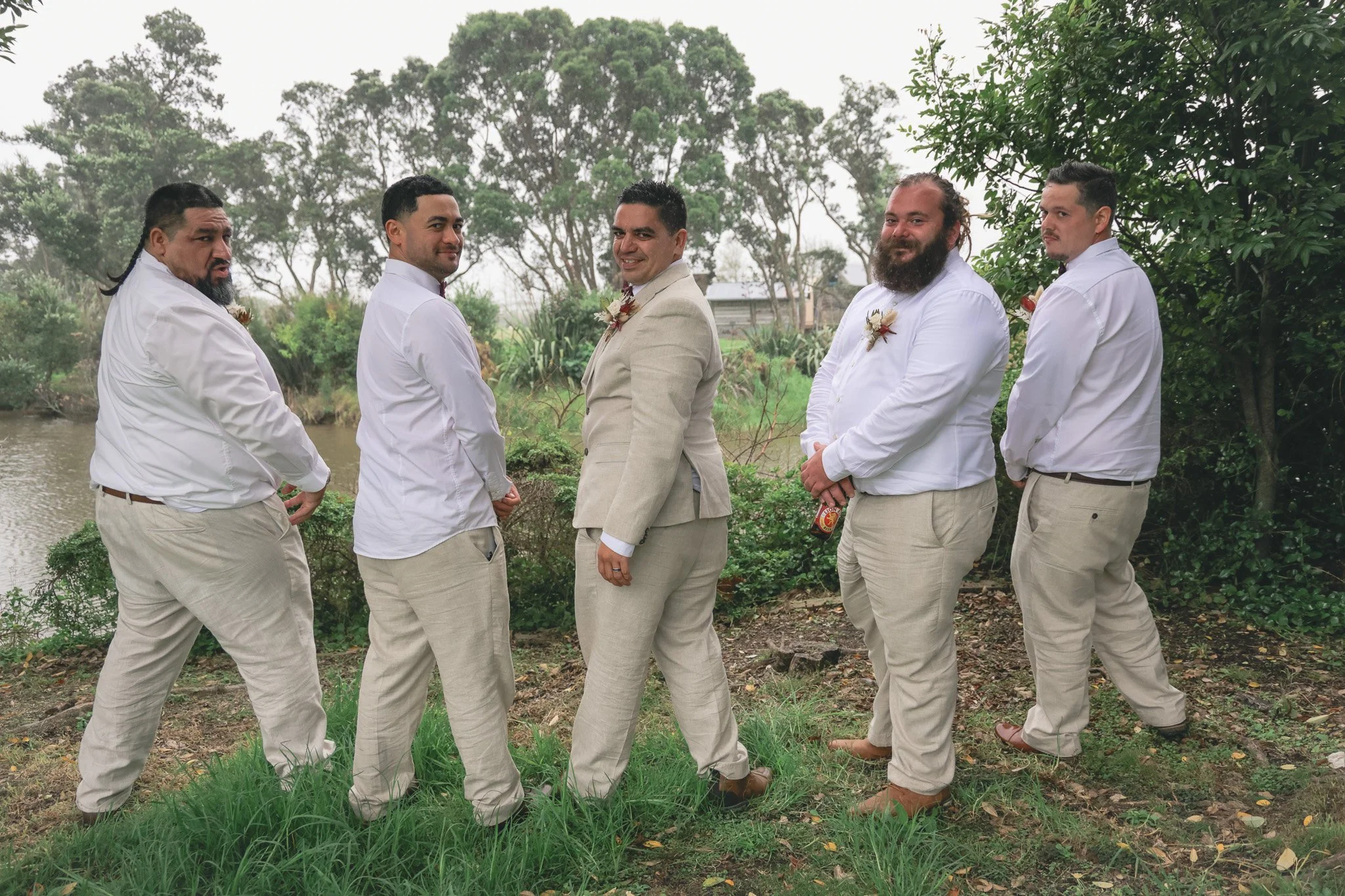 Group of five men dressed in beige and white suits standing outdoors near water with trees in the background, posing for a photo during a daytime event.