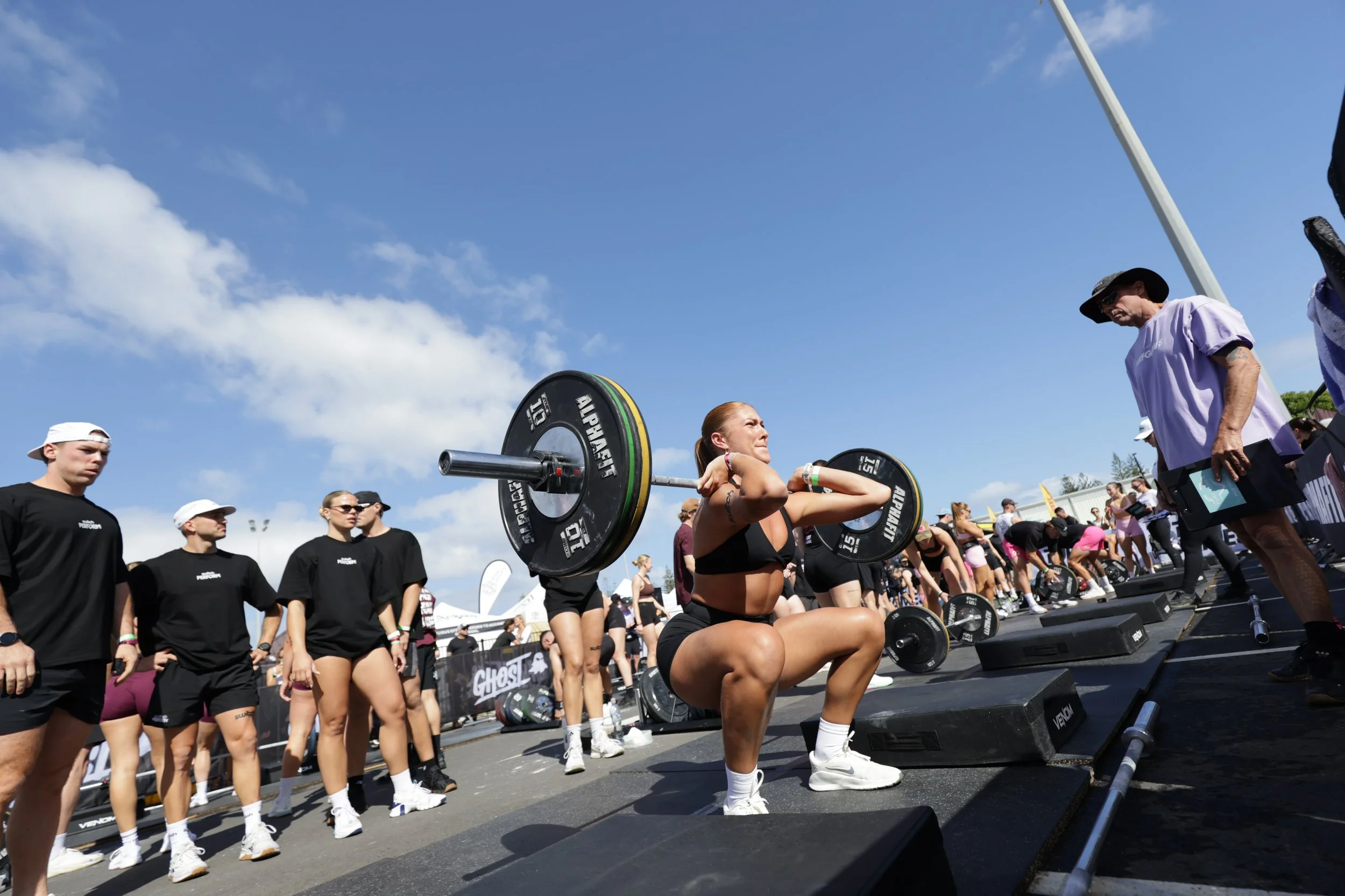 A woman performing a squat exercise with a barbell at an outdoor fitness event, surrounded by people and a clear blue sky.