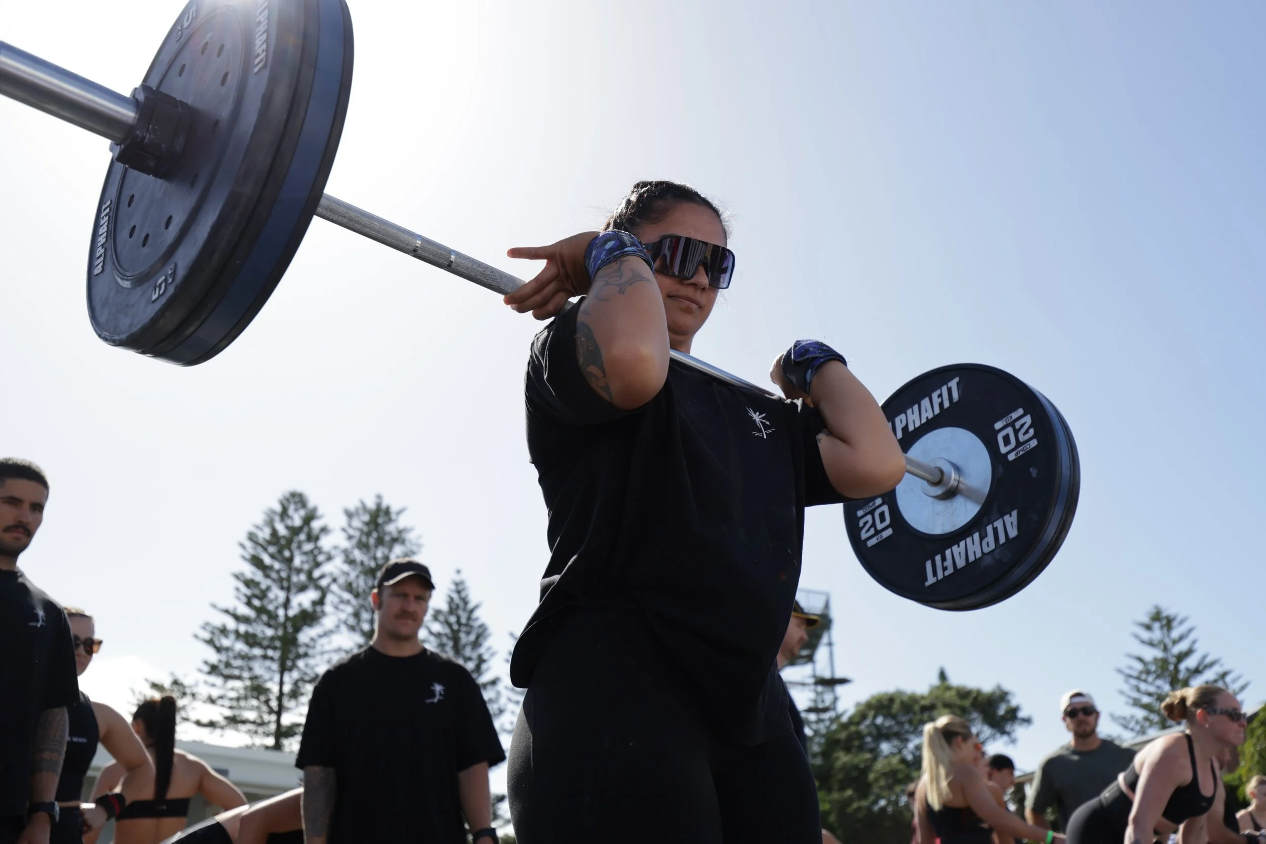 A woman wearing sunglasses lifting a barbell with weights during a fitness event, with multiple people gathered around outdoors under a clear sky.