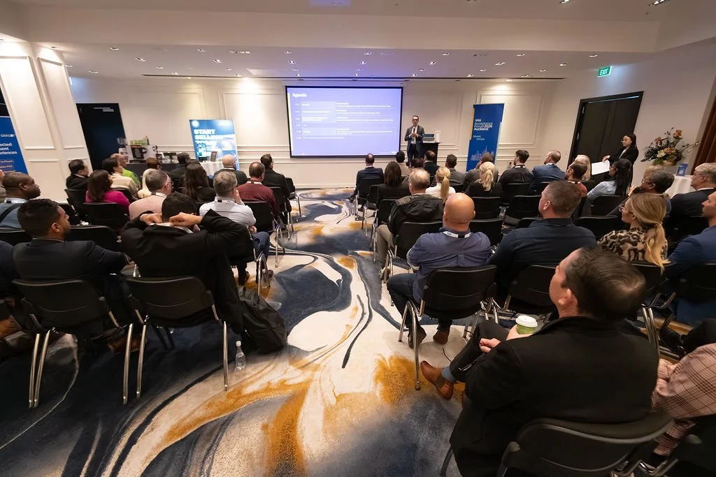 A professional conference with a speaker presenting to an audience in a hotel conference room, with a large screen displaying presentation slides.