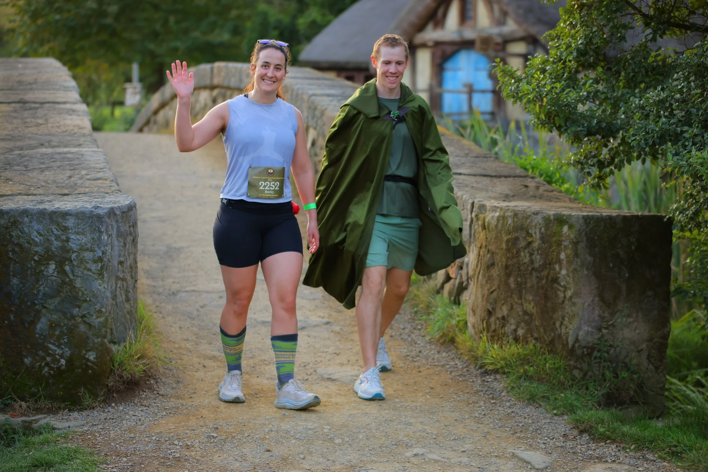 Two young people, a woman and a man, walking on a dirt path outdoors near a stone bridge and trees. The woman is smiling and waving at the camera, wearing a light blue sleeveless top, black shorts, and colorful socks. The man is smiling and looking d