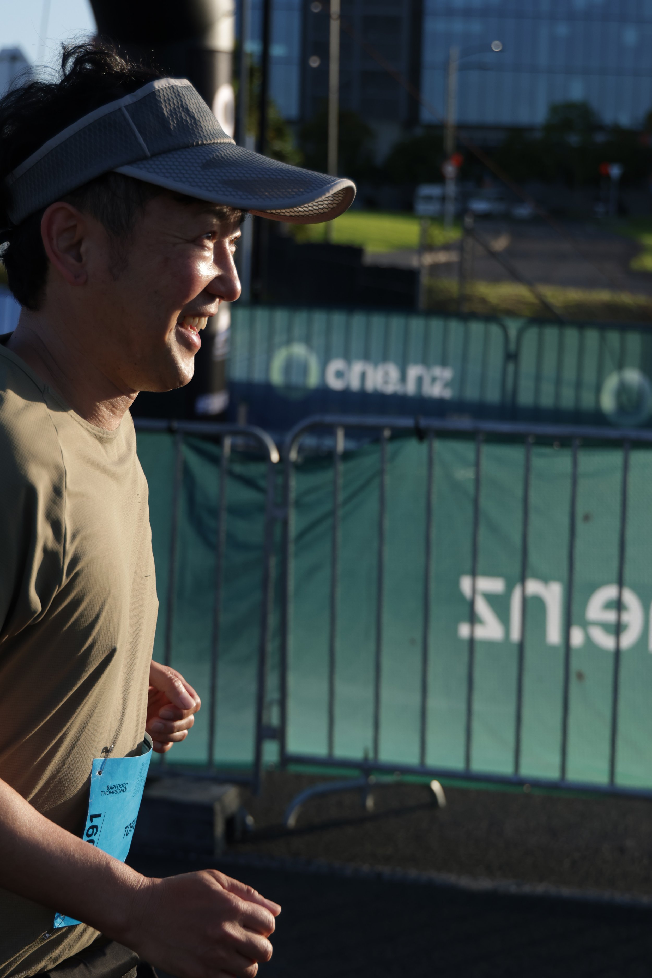 A woman in a beige shirt and a gray visor cap is running outdoors during the daytime. She has a smile on her face and a race bib attached to her shirt. The background includes a green fence with text, a parking lot, and buildings in the distance.
