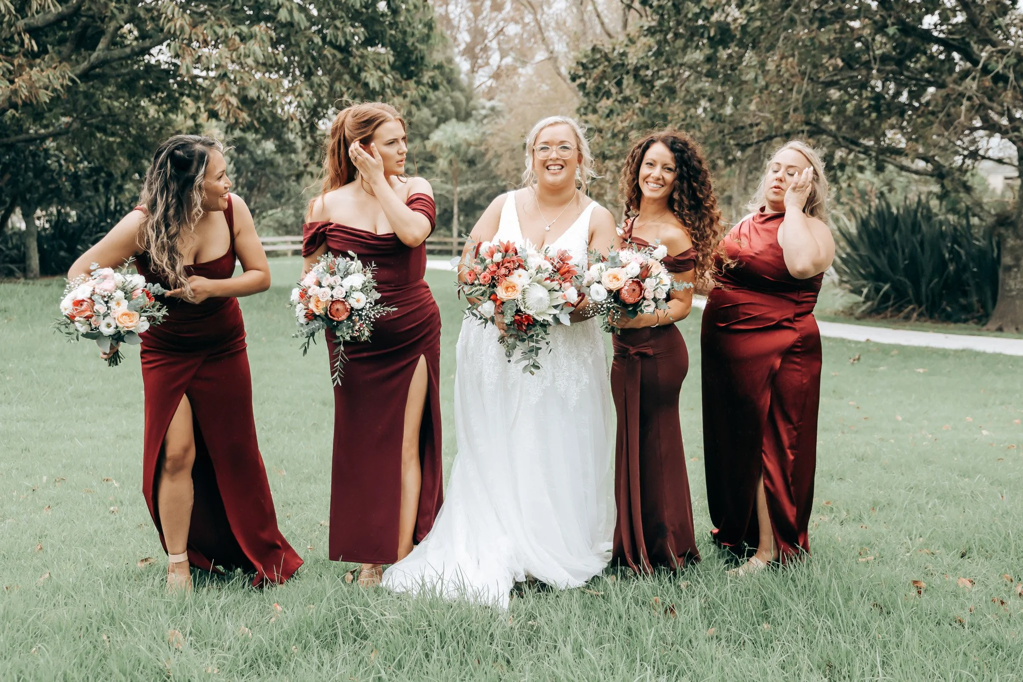 A bride and four bridesmaids standing outdoors on a grassy area surrounded by trees. The bride is in a white wedding dress holding a bouquet, and the bridesmaids are in matching burgundy dresses holding smaller bouquets.