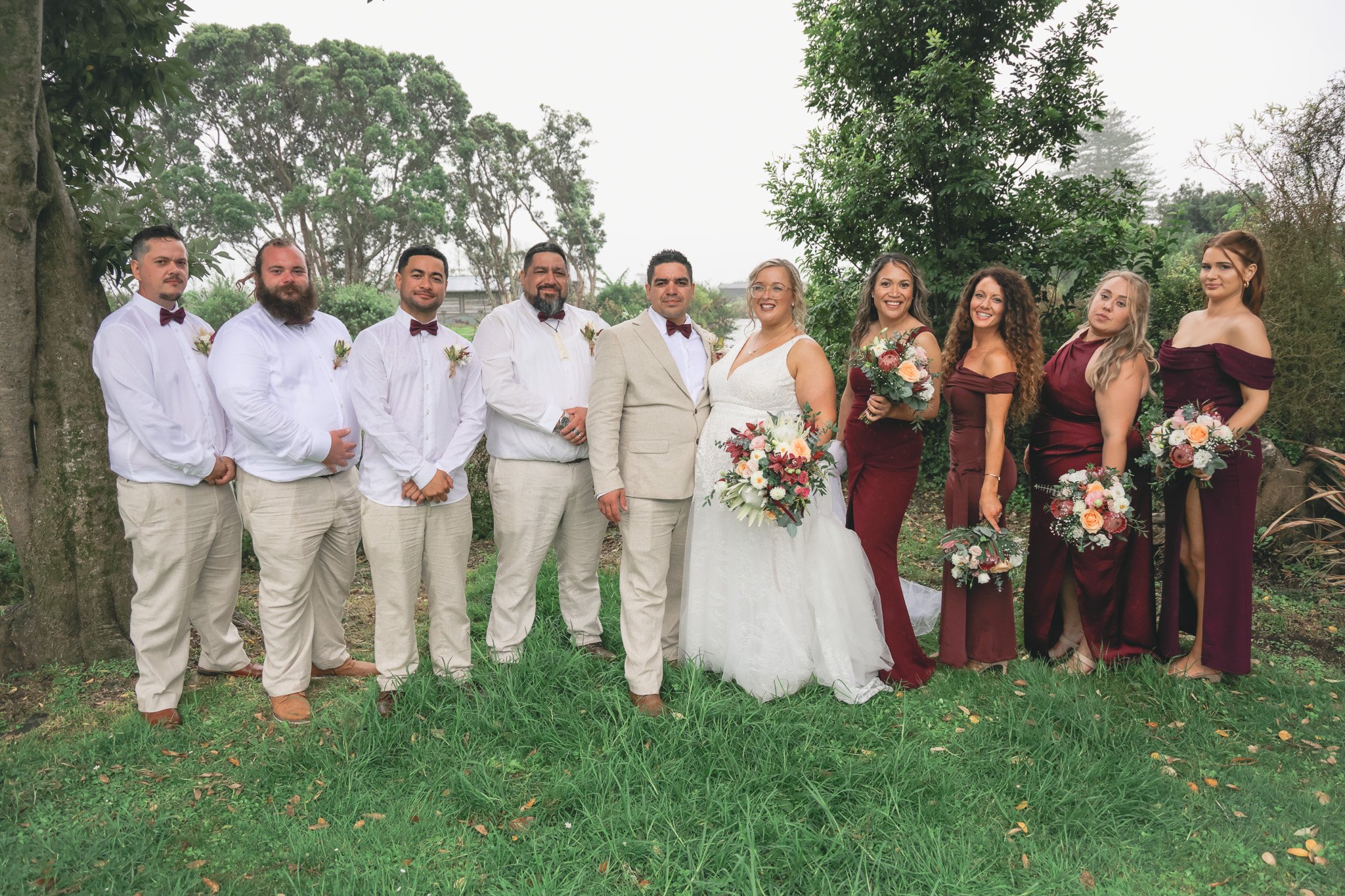 Group of wedding party members standing outdoors under trees, including bride and groom, bridesmaids, and groomsmen, all dressed in formal attire with bouquets and boutonnieres.