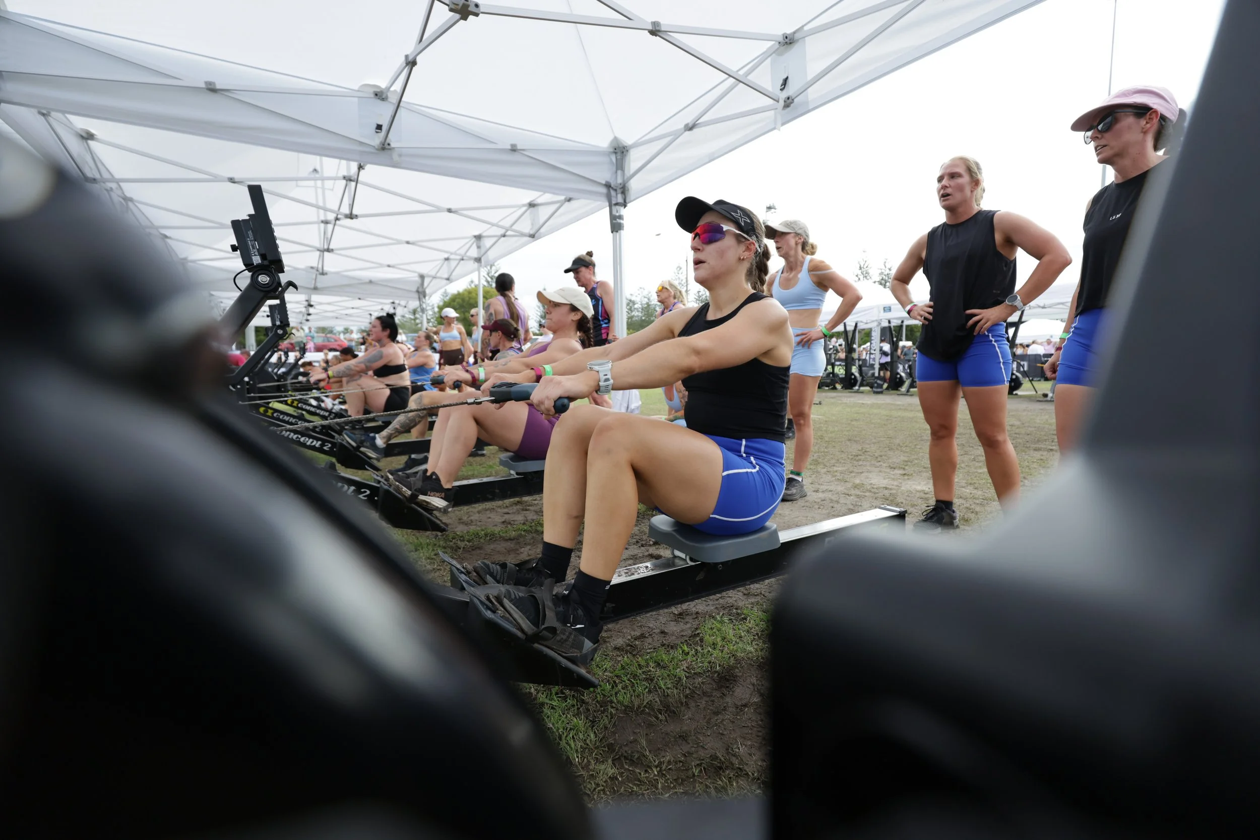 Women participating in an outdoor rowing event, with some sitting on rowing machines under a white tent and others standing nearby, during daytime with cloudy sky.