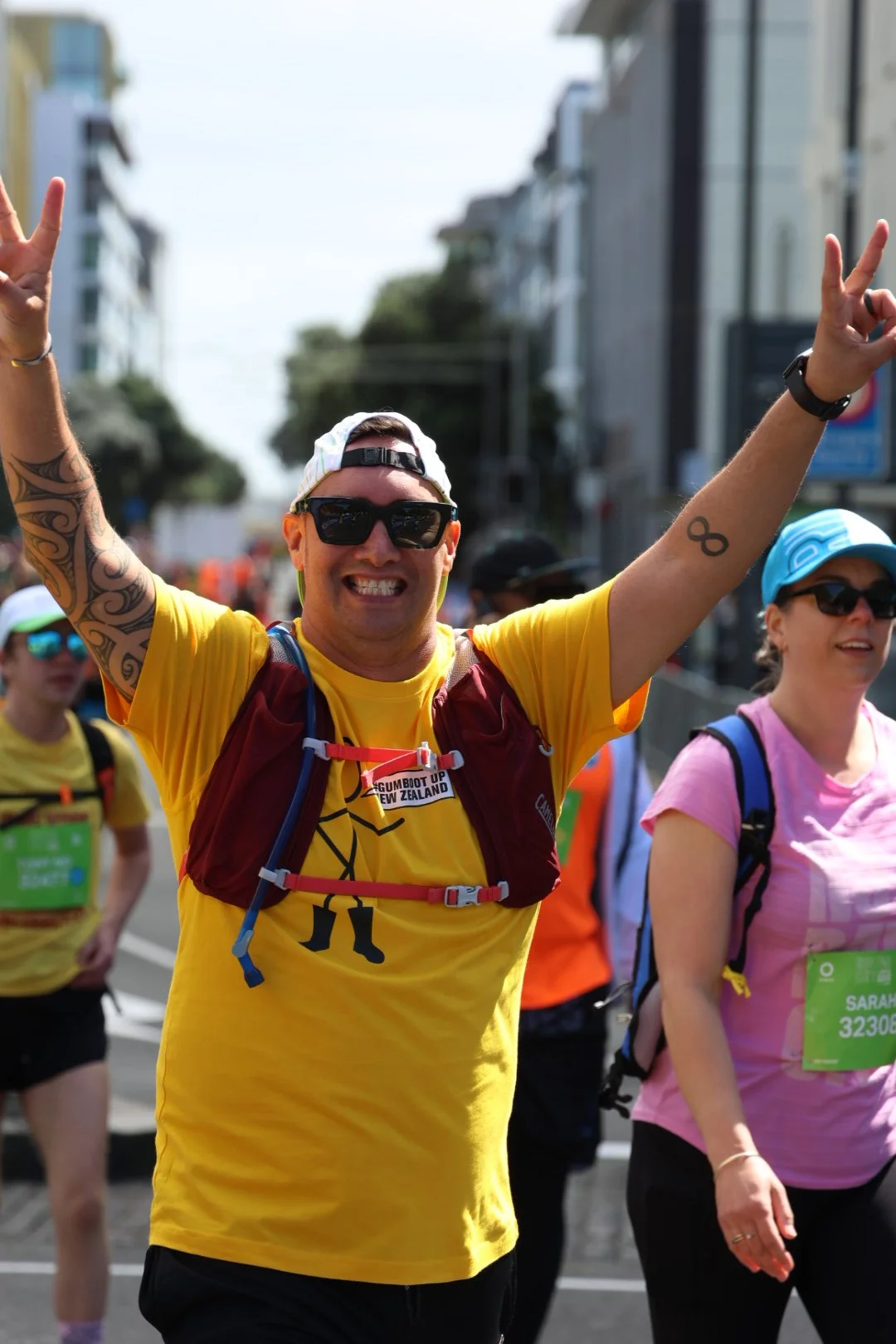 A man in a yellow shirt with tattoos on his arm, sunglasses, and a baseball cap, smiling and making peace signs during a marathon or running event on a city street.