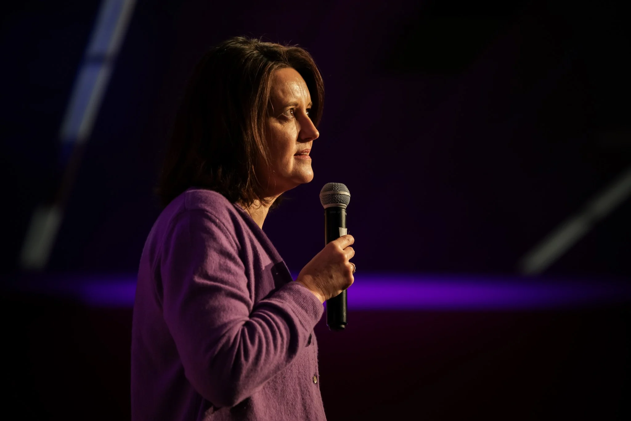 A woman with shoulder-length brown hair wearing a purple blazer is holding a microphone and speaking on stage with a dark background.