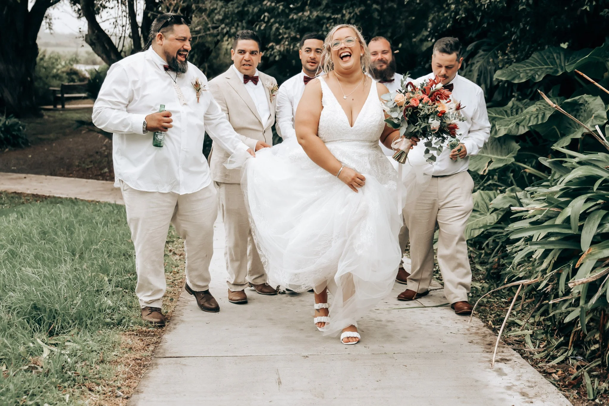 A bride in a white wedding dress walking on a garden path, smiling, holding a bouquet, surrounded by five men in white shirts and tan pants with bow ties, outdoors under trees.