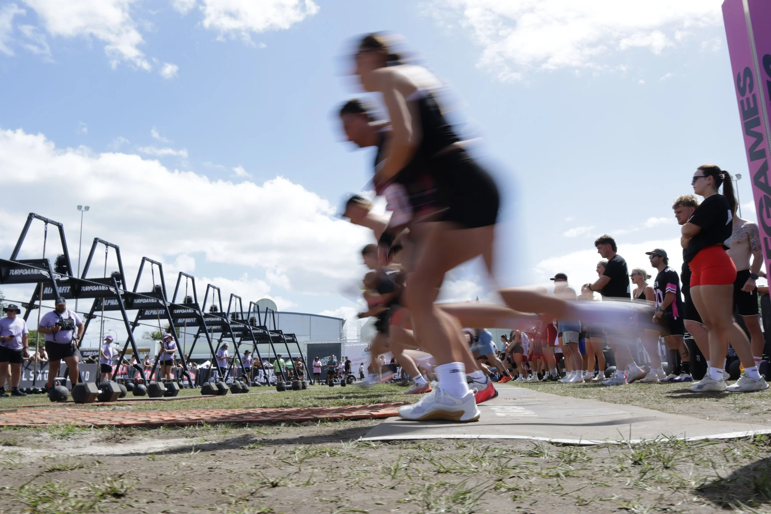 Blurred runners participating in a race at an outdoor event under a partly cloudy sky, with spectators and event equipment in the background.