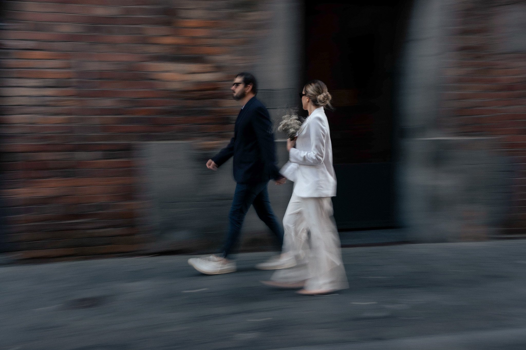 A man in a dark suit and a woman in a white suit walk together along a city sidewalk near a brick wall.