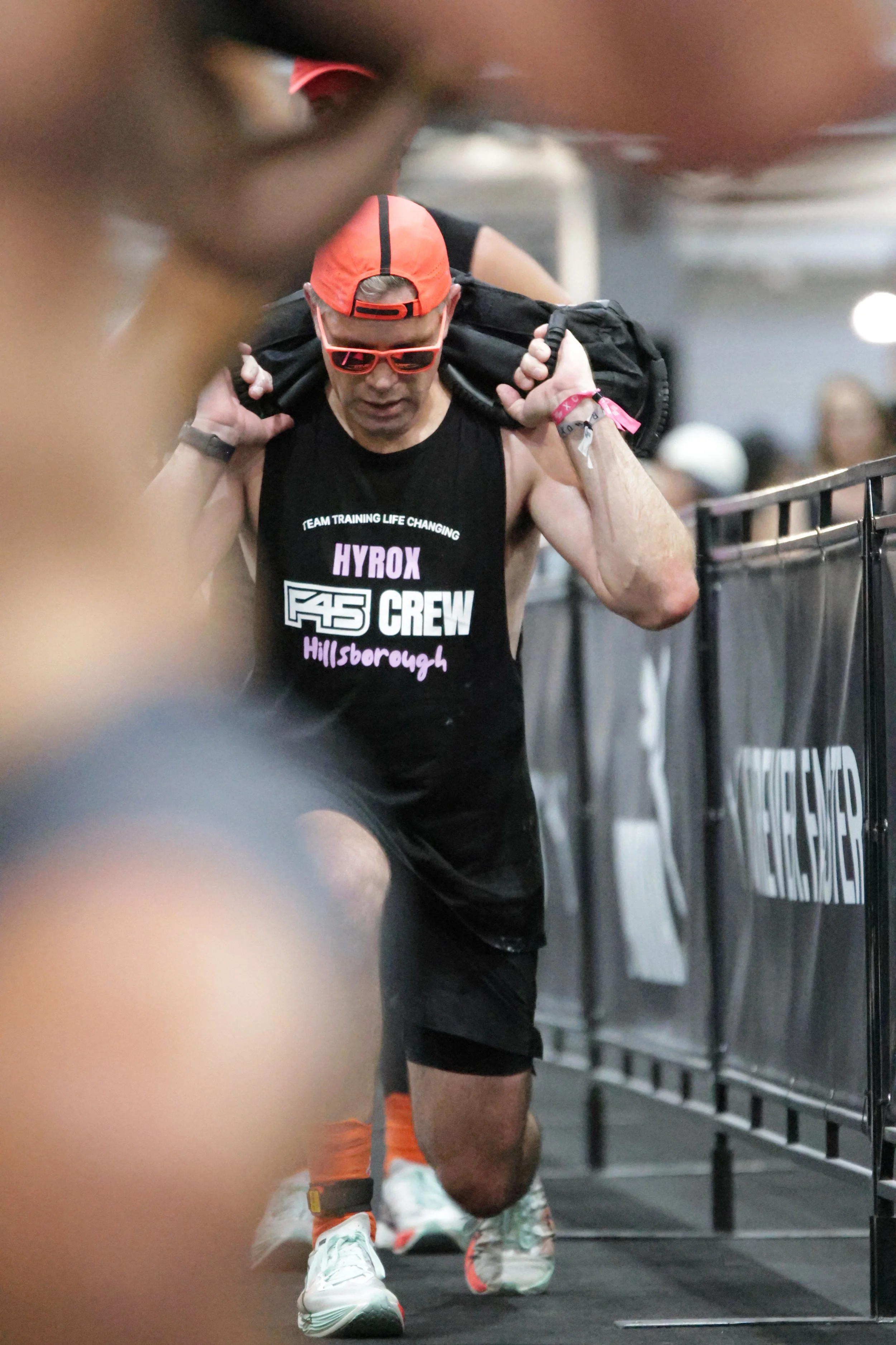 A male athlete wearing a black sleeveless shirt with race team logos, red cap worn backwards, red sunglasses, black shorts, and white athletic shoes with orange accents, carrying a black gym bag on his shoulders, walking in an indoor gym or sports fa