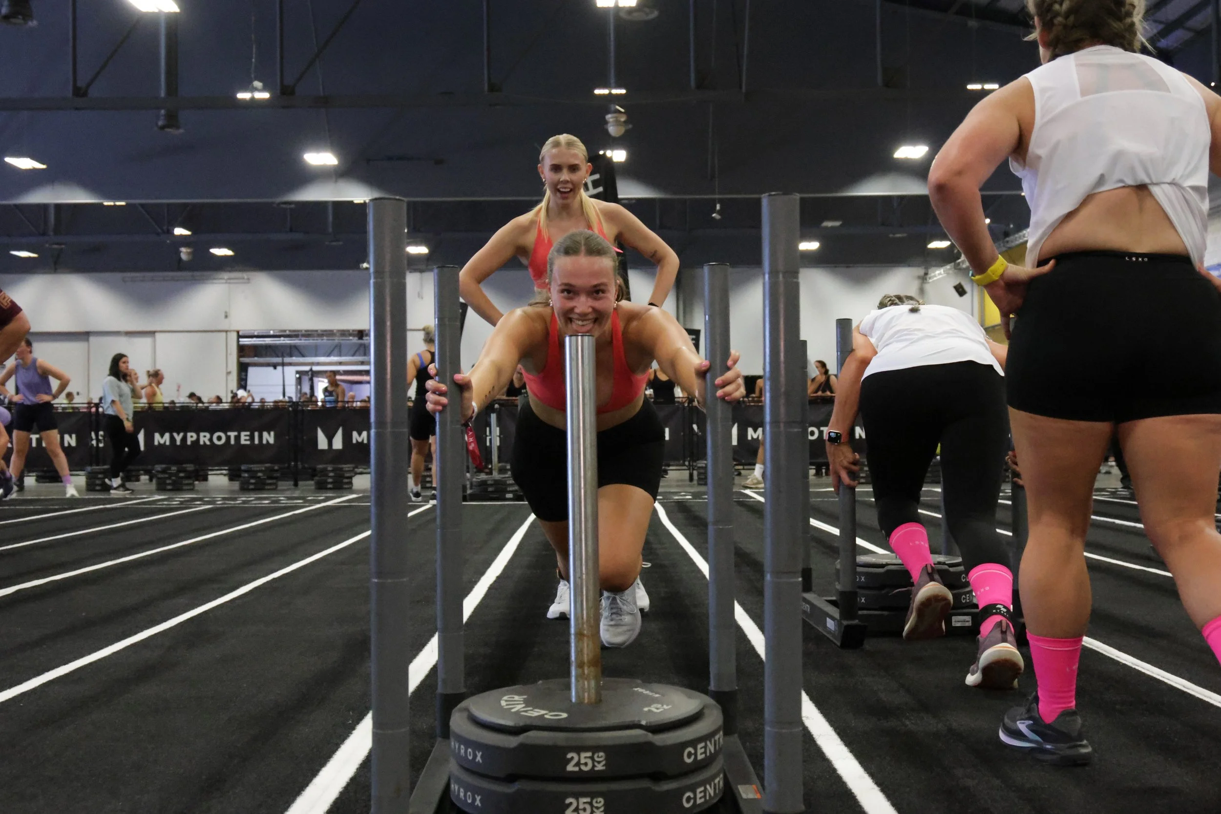 Two women are participating in a fitness event in an indoor gym. One woman is smiling and pushing a weighted sled, while the other woman is observing and standing nearby. There are other participants and spectators in the background, with large black
