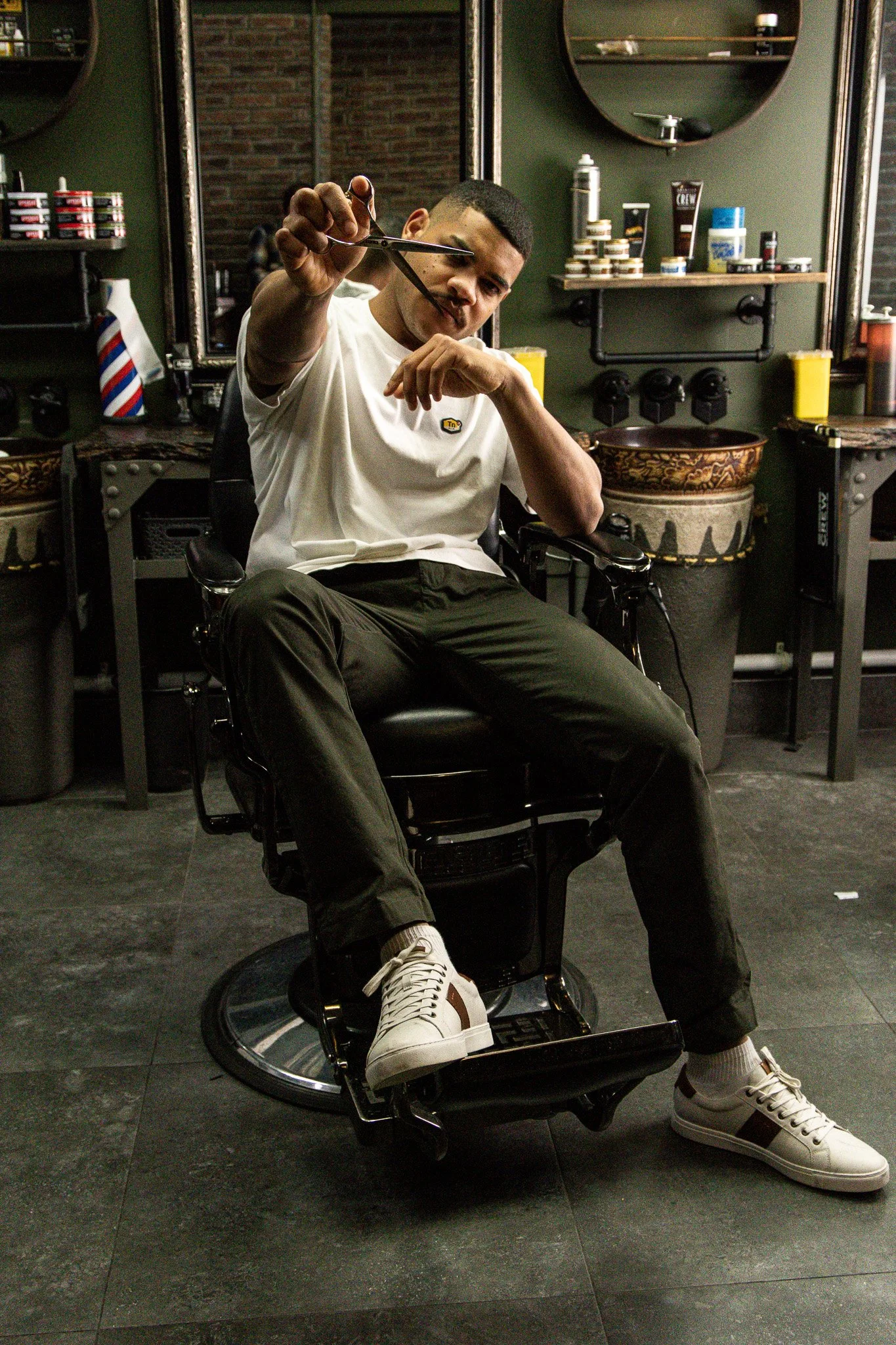Man seated in a barber shop chair, holding scissors, with grooming products on shelves and a mirror behind him.