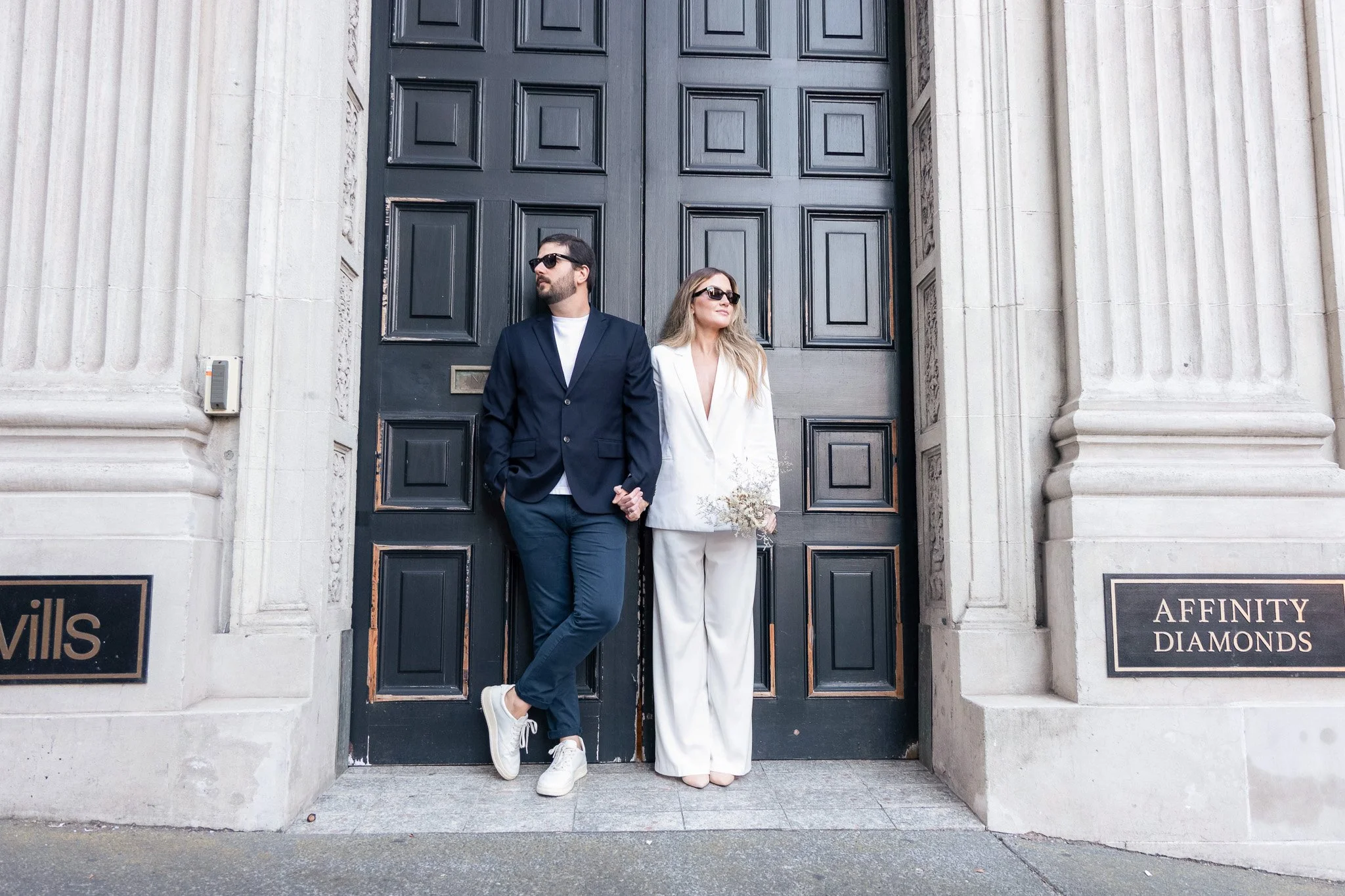 A man and woman leaning against a large black door with decorative panels, in front of a stone building with signs reading "A pretty" and "Affinity Diamonds." The man has dark hair and beard, wearing sunglasses, a navy blazer, white T-shirt, dark pan