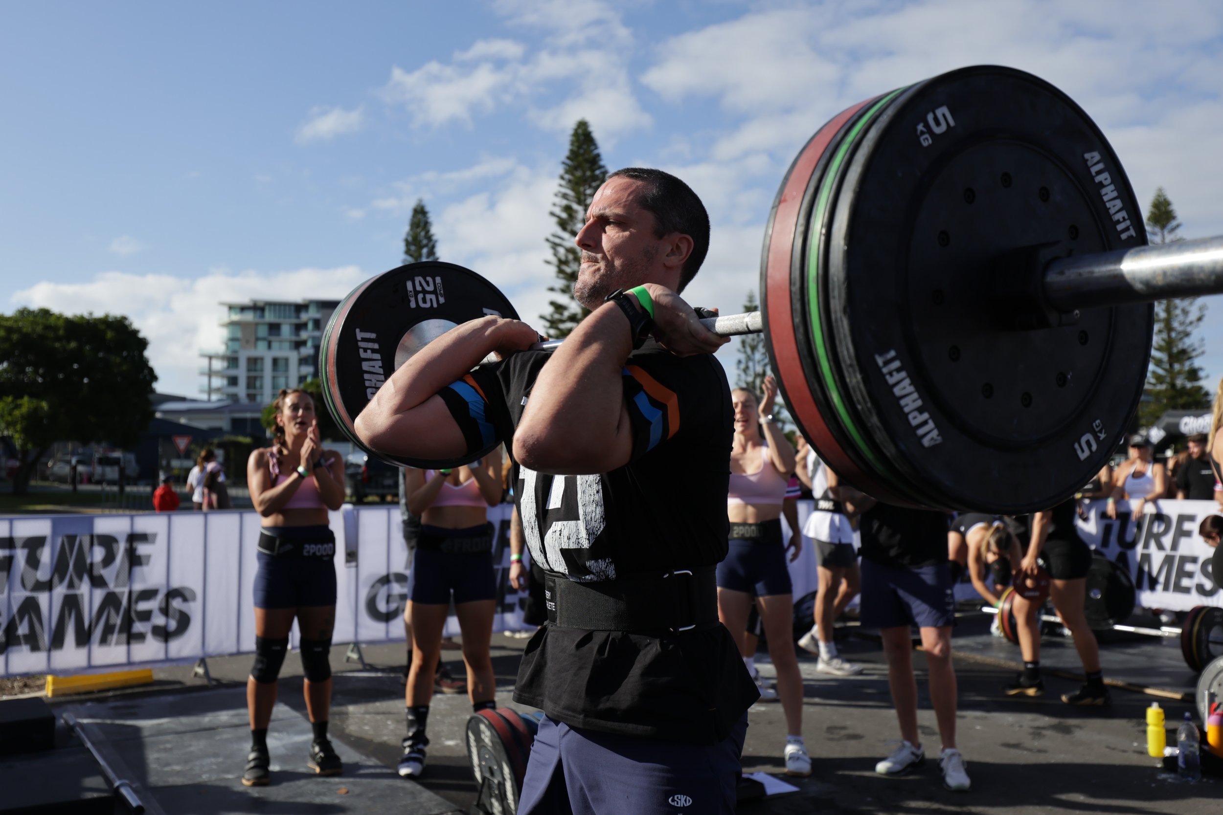 A man is lifting a barbell with weight plates during a weightlifting competition outdoors. Several women are standing behind him, clapping and cheering. There are trees, a building, and a fence with banners in the background on a sunny day.