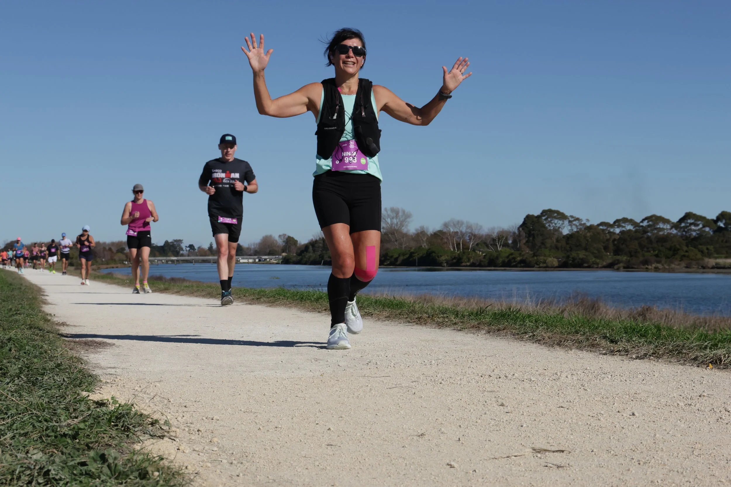 A woman with short dark hair, sunglasses, and black athletic clothing runs on a dirt path beside a body of water, participating in a race with other runners behind her. She is smiling and waving at the camera. The sky is clear and blue, and there are