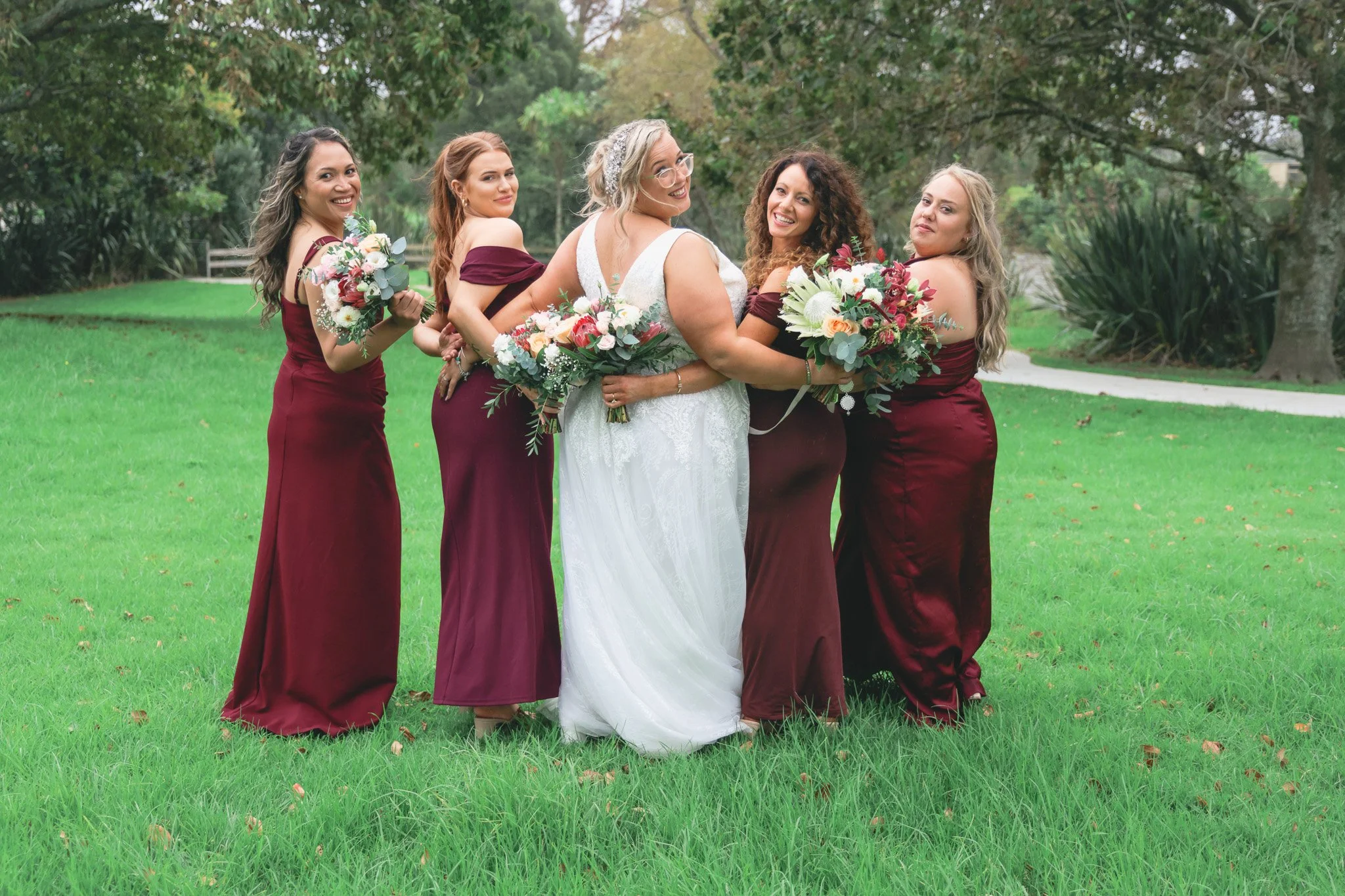 A bride in a white wedding dress and five bridesmaids in burgundy dresses standing outdoors on green grass, holding bouquets of flowers.
