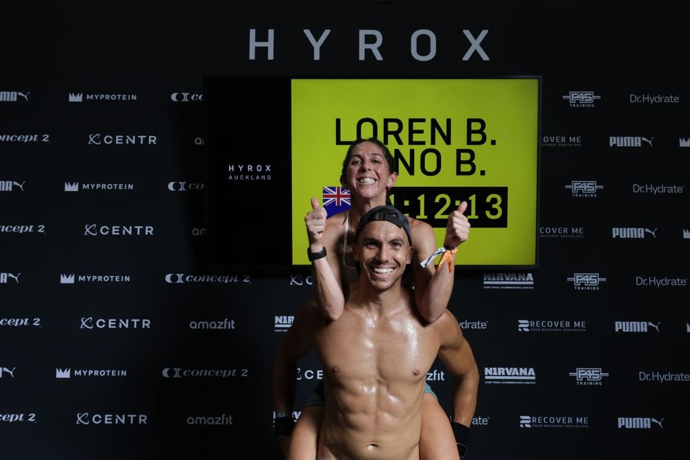 A woman sitting on a man's shoulders, both smiling and giving thumbs up. They are at the HYROX finish line in Auckland, with a large yellow screen behind them displaying their names, a flag, and the time 1:12:13.