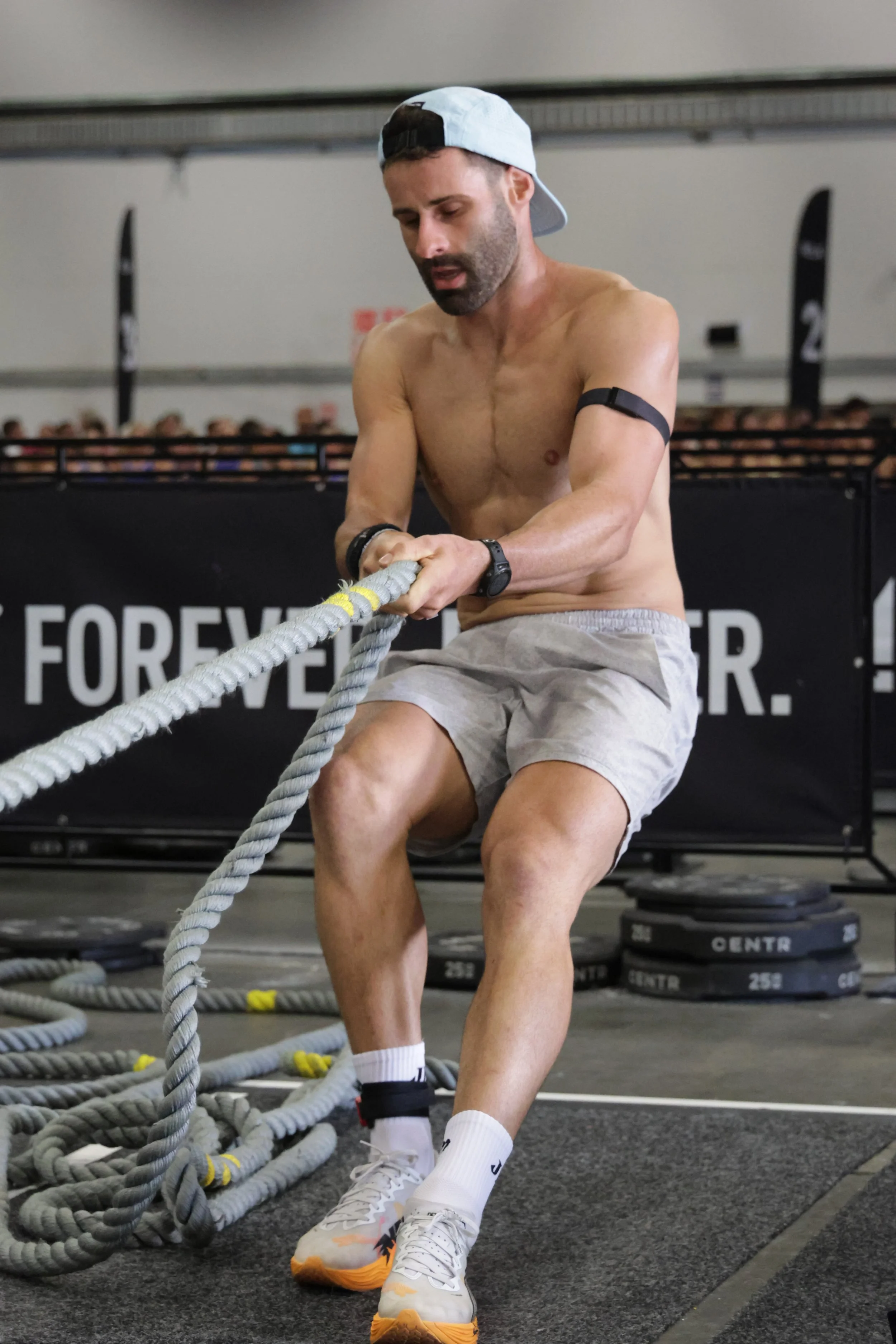 A shirtless man wearing a backwards baseball cap, shorts, and sneakers, working out with battle ropes in a gym.