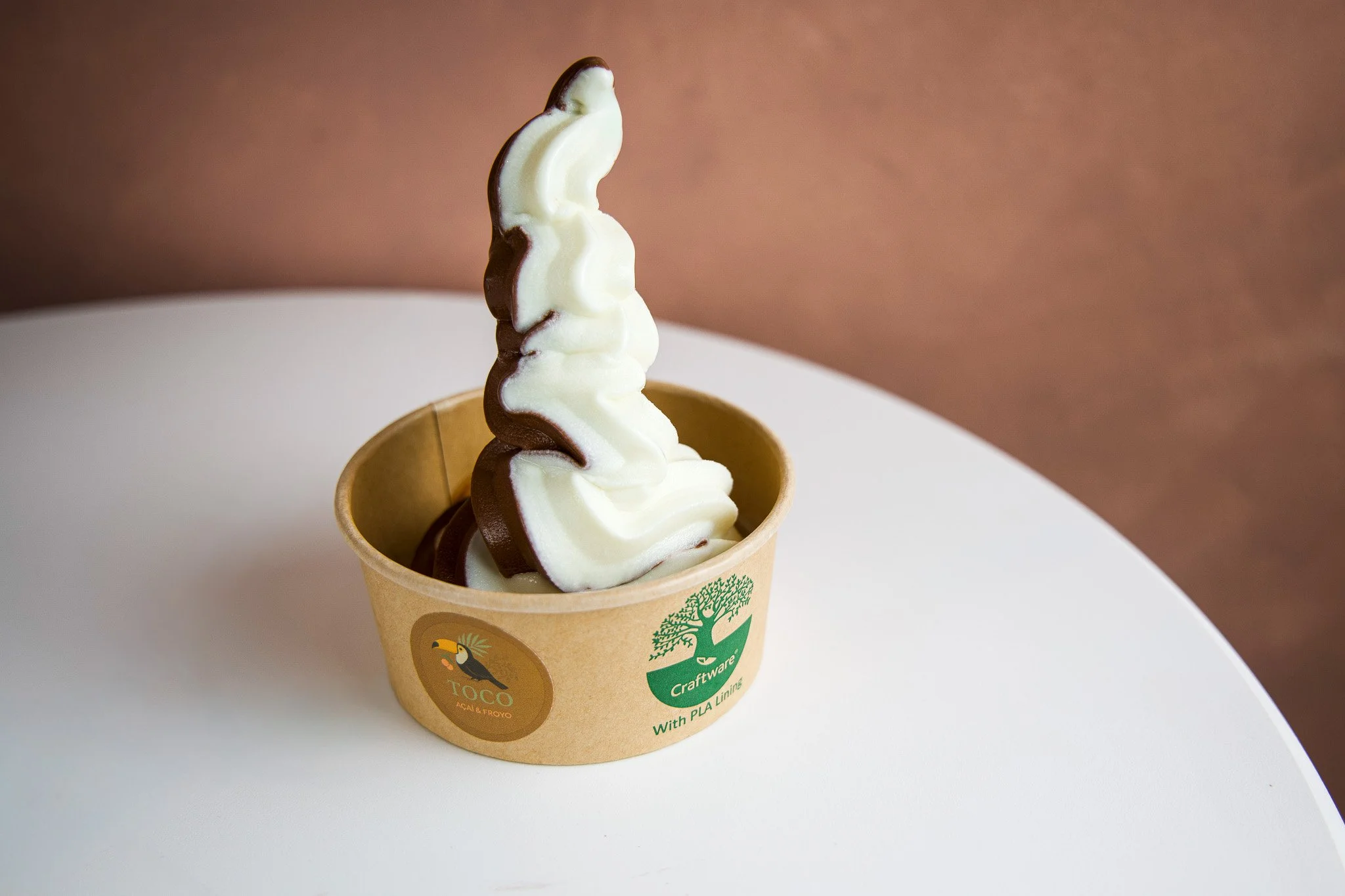 Soft serve ice cream with chocolate and vanilla swirls in a paper cup with branding, placed on a white surface against a brown background.