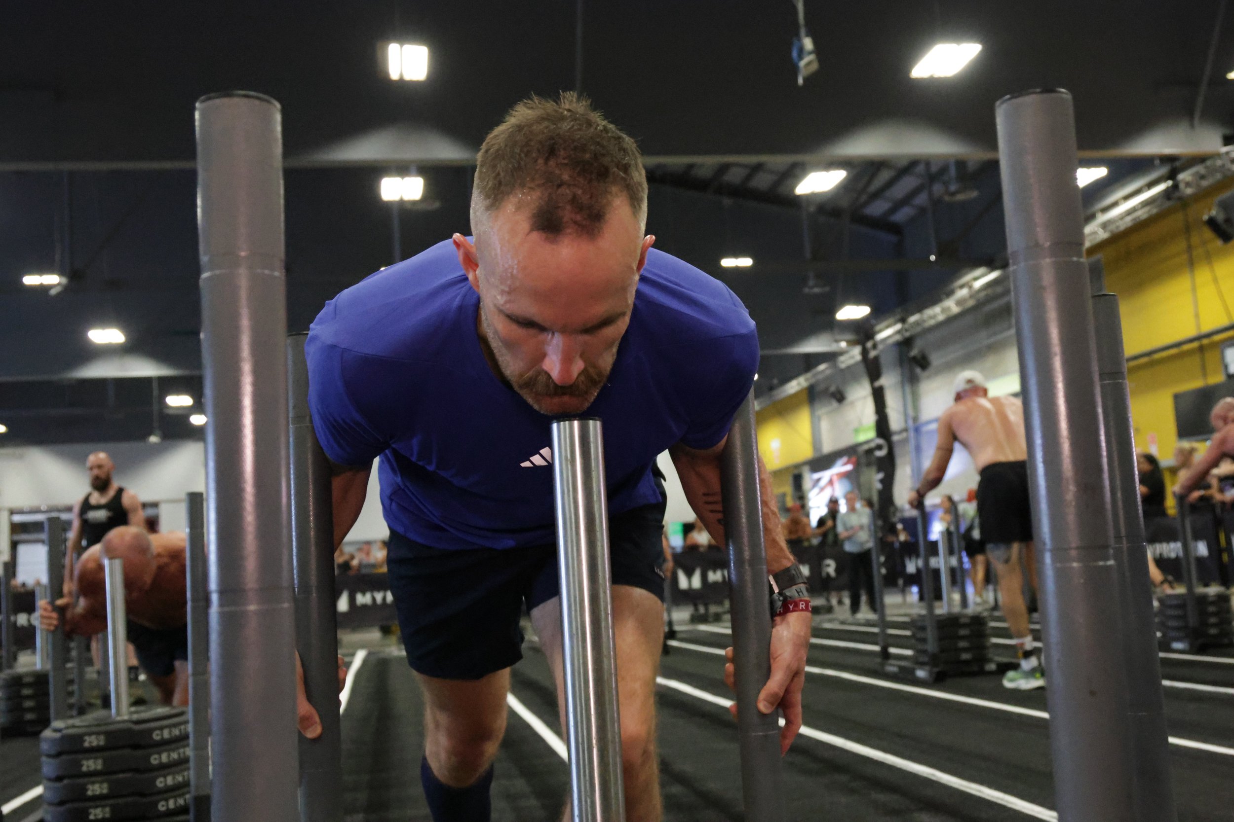 Man in a blue shirt doing push-ups on a black gym floor with people working out in the background at a fitness center.