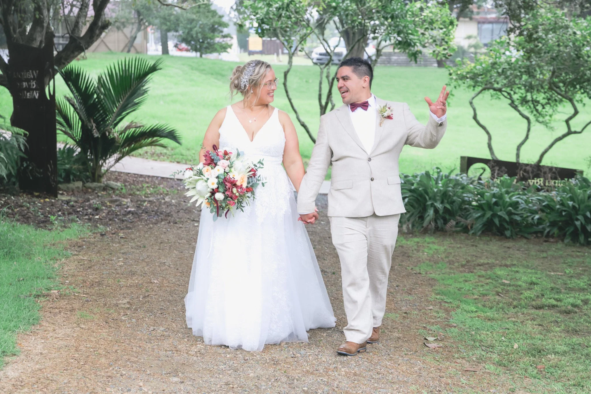 A bride and groom holding hands and smiling at each other outdoors in a park, dressed in wedding attire, with the groom making a peace sign with his other hand.