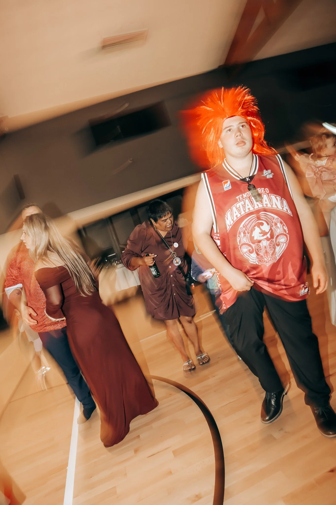 People at a party or gathering, including a young man with bright orange, spiky hair wearing a red sports jersey. Others are seen in the background, some talking or holding drinks, in a room with wooden flooring and beige walls.