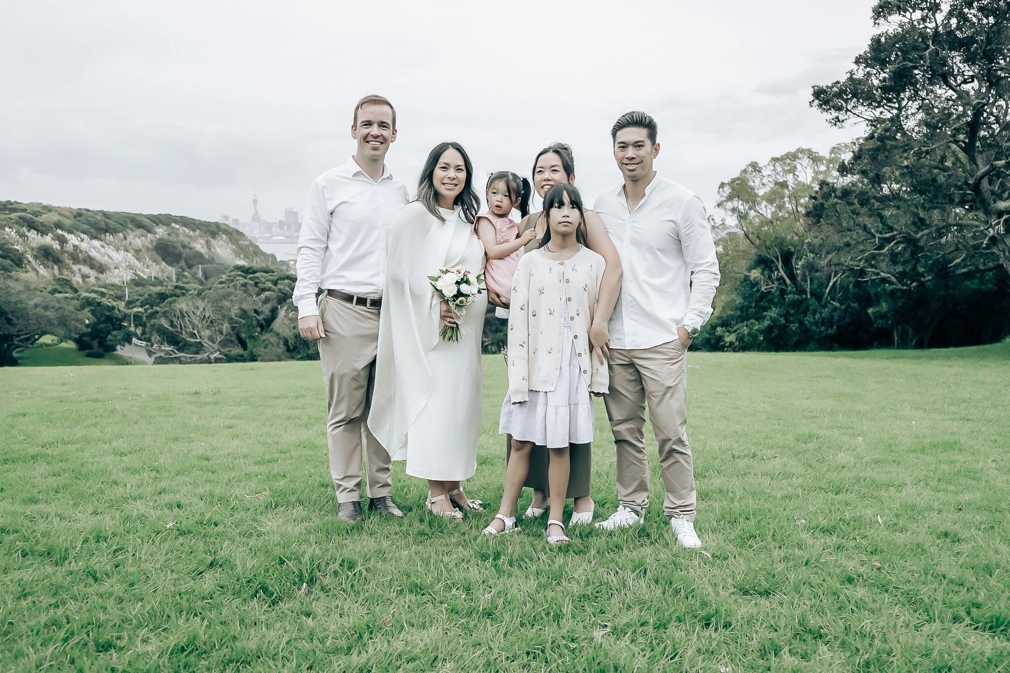 A diverse group of six people, including two children, standing outdoors on a grassy field with trees and a city skyline in the background. They are smiling and dressed in light-colored clothing.