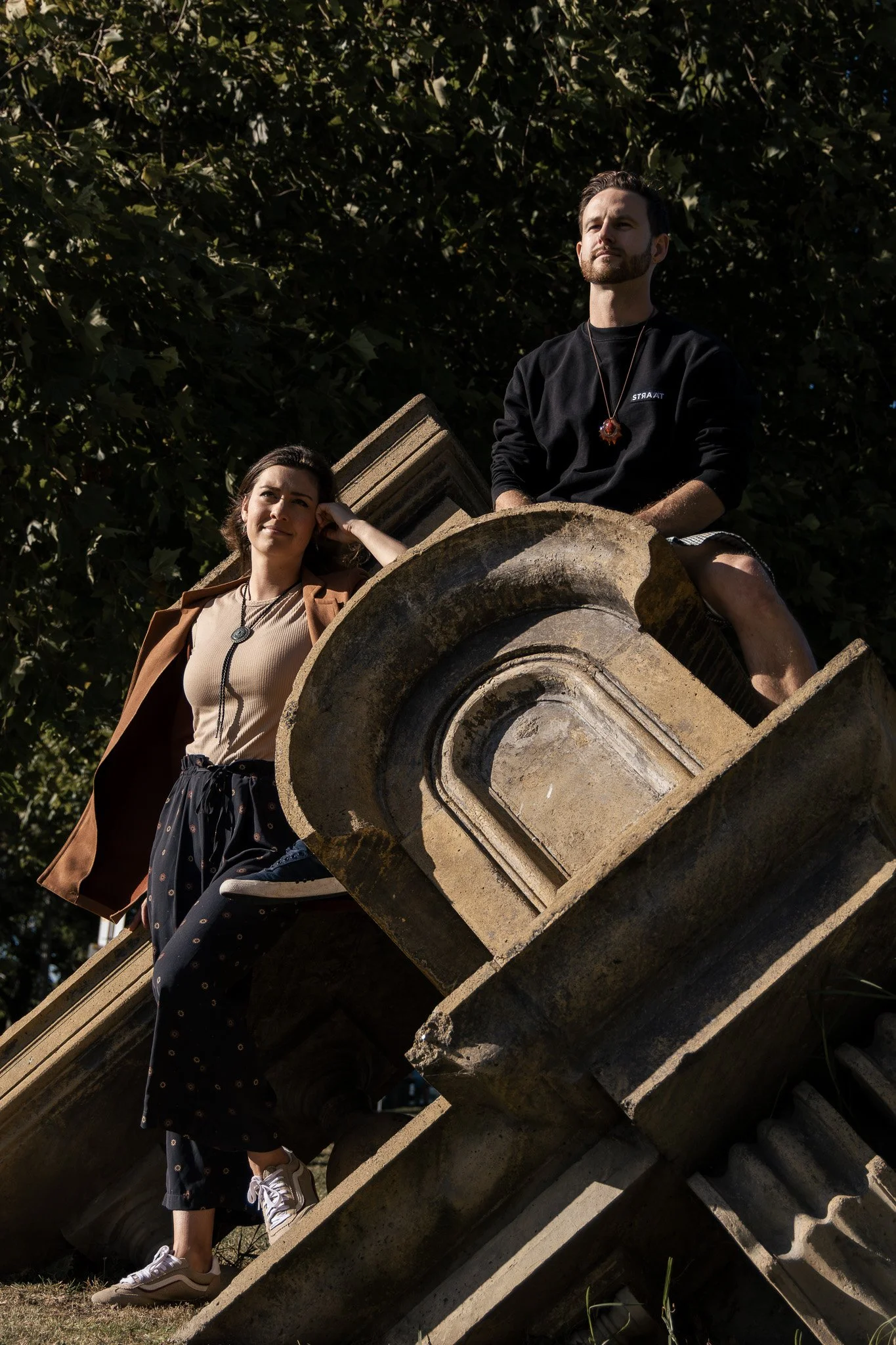 Two young adults, a woman with dark hair and a man with a beard, sit on a large, decorative stone structure outdoors during daytime. The woman is leaning against the structure, looking into the distance, while the man is sitting with his legs crossed