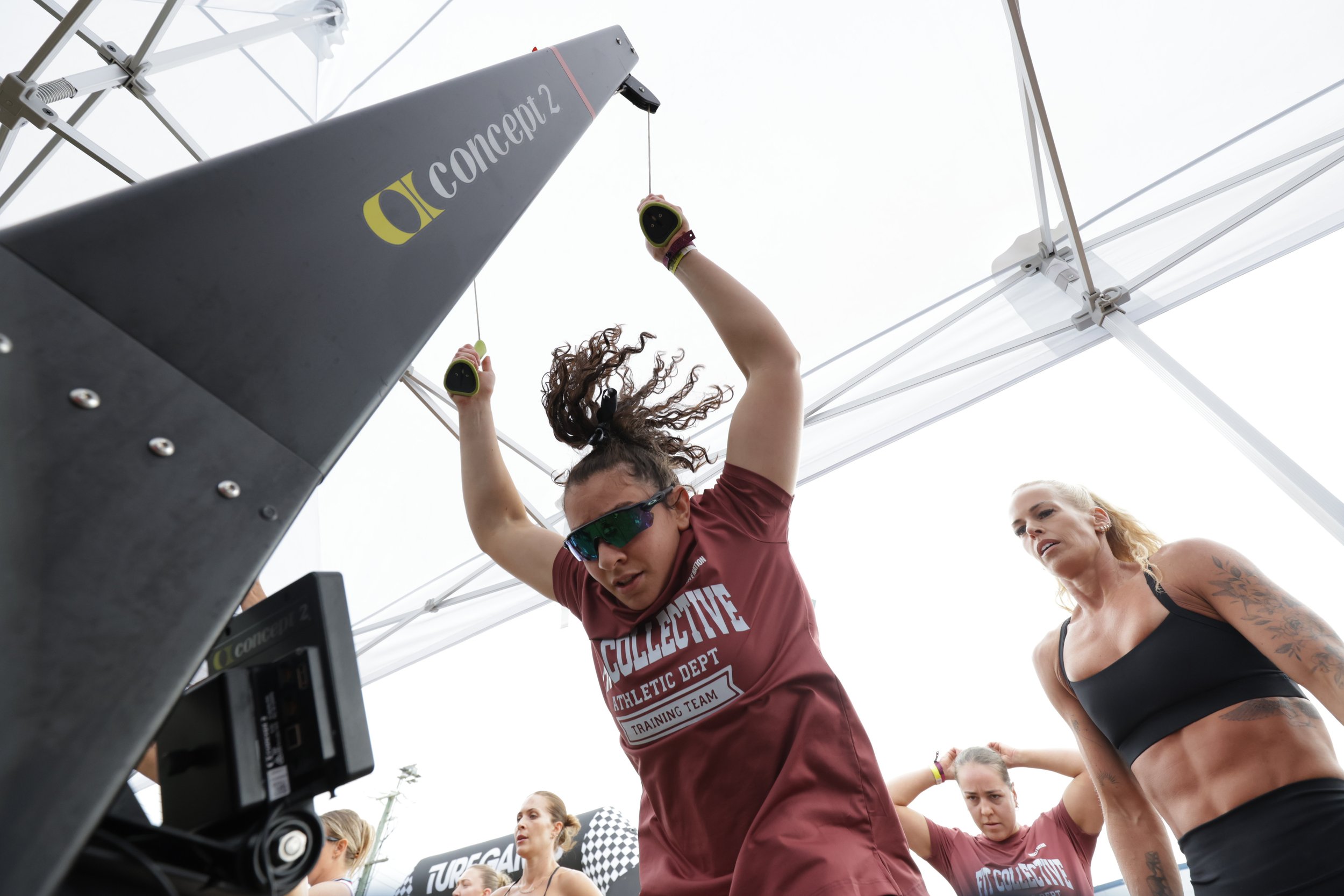 A group of women participating in an outdoor fitness event, with one woman wearing sunglasses and a maroon shirt, actively lifting a barbell with weights while others observe or wait their turn.