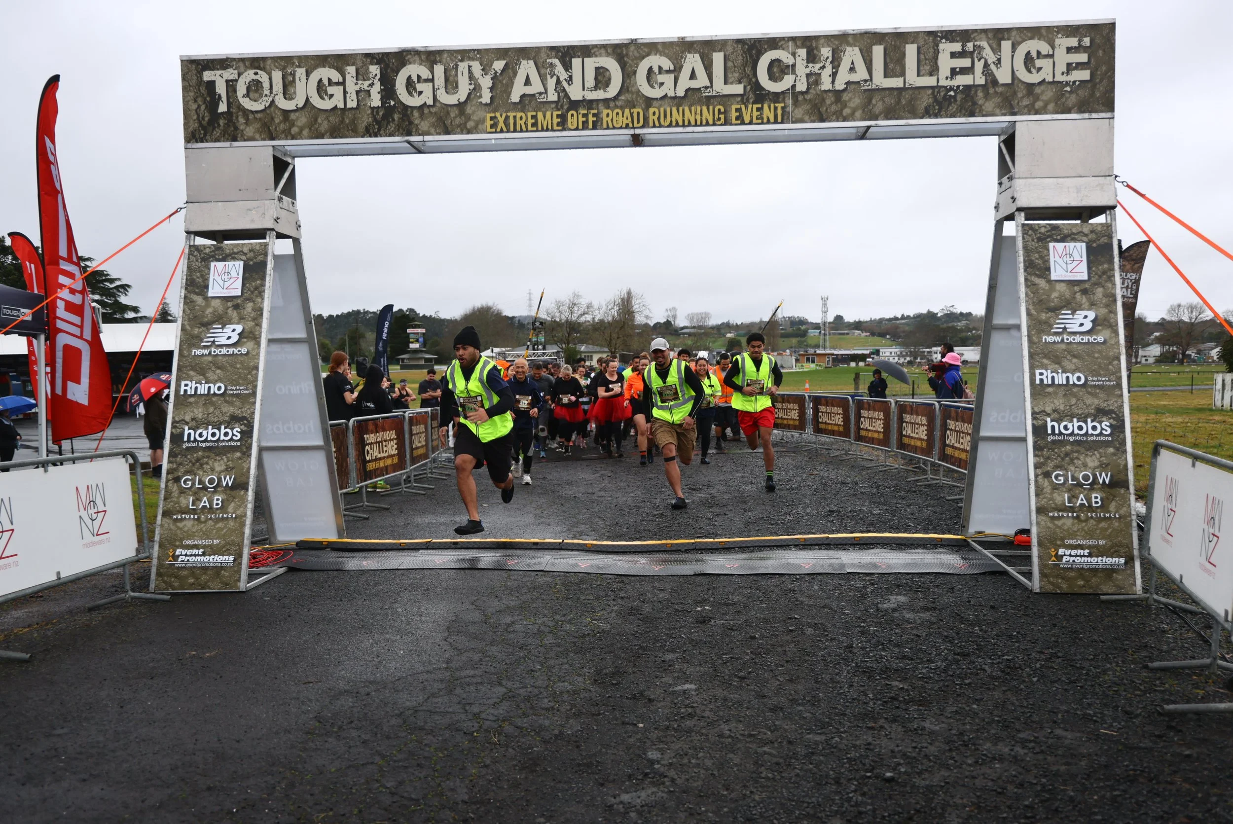 Participants running in an outdoor race event called 'Tough Guy and Gal Challenge' under a large banner, on a cloudy day with a grassy field and trees in the background.