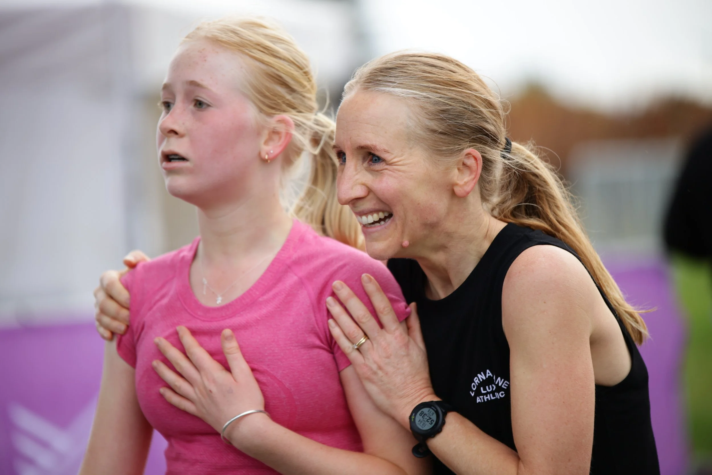 A woman and a young girl are smiling and embracing during an outdoor activity, possibly sports or exercise, with a purple and green background.