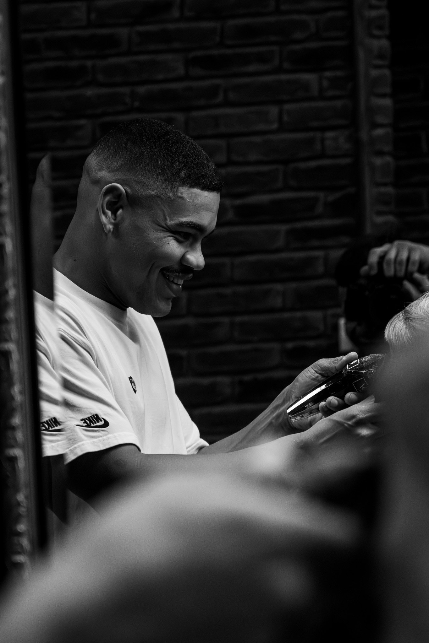 A young man with short hair and a smile, wearing a white Nike shirt, looking at a phone while sitting indoors with a brick wall in the background.