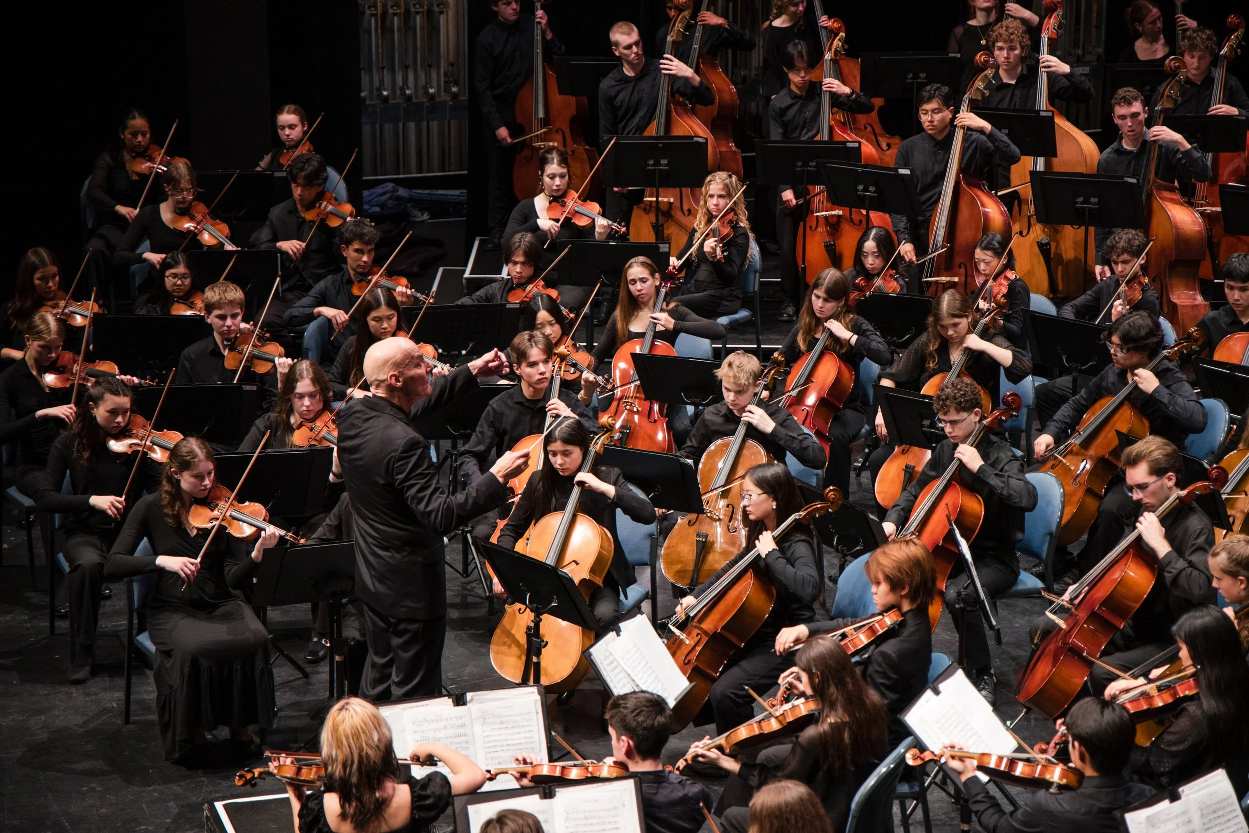 An orchestra of young musicians performing on stage, playing violins, cellos, and double basses, led by a conductor.