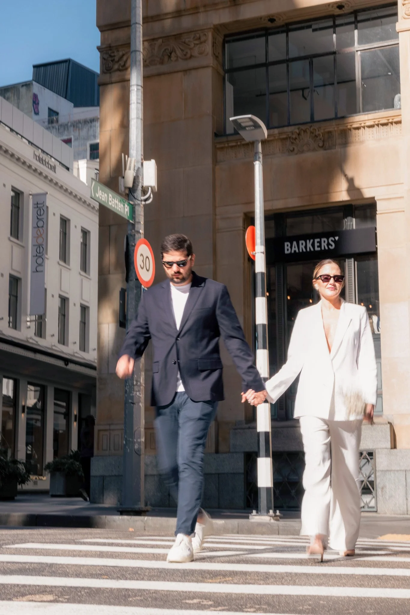 A man and a woman holding hands and crossing the street on a crosswalk in an urban area, with buildings and street signs visible in the background.