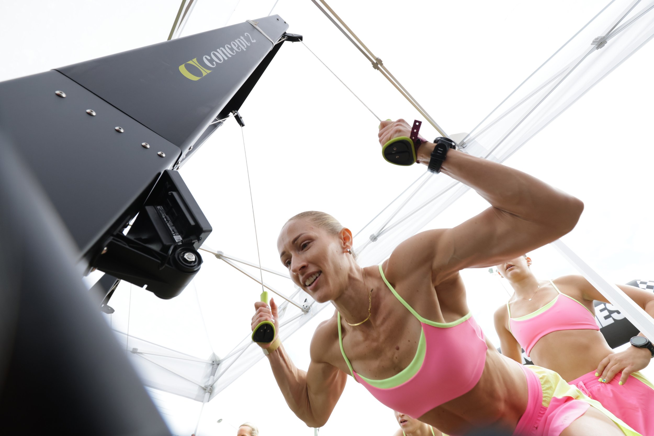 A woman in bright pink athletic wear is participating in a fitness competition, using resistance bands while a person above her adjusts equipment. The scene takes place outdoors under a white tent.