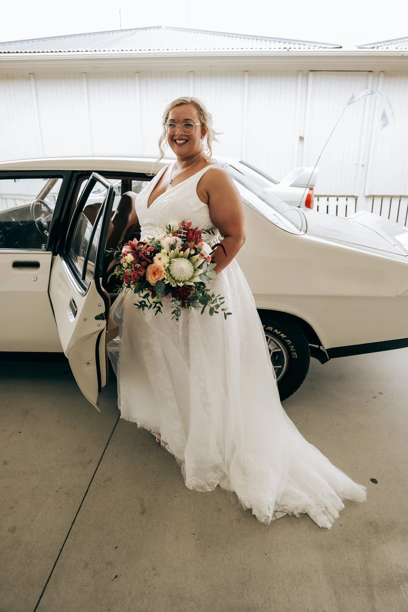 A smiling woman in a white wedding dress holding a bouquet of flowers, standing next to an open car door in a garage or similar indoor space.