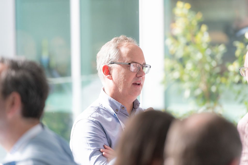 A group of people in a meeting room with a man wearing glasses and a light blue shirt, standing near a window and speaking.