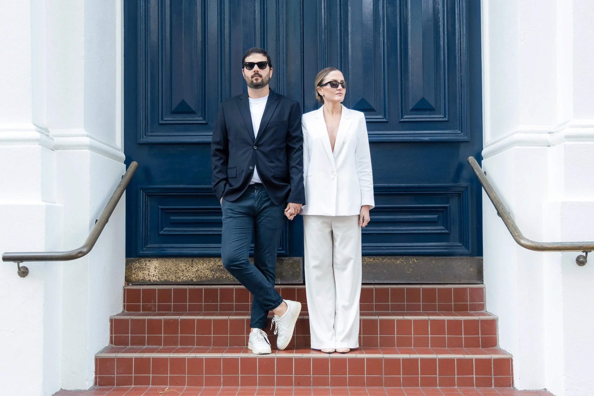 A man and woman standing on red tiled steps in front of a dark blue door, holding hands and looking away from the camera, both wearing sunglasses. The man is dressed in a black blazer, white shirt, and jeans, while the woman is in a white suit with a