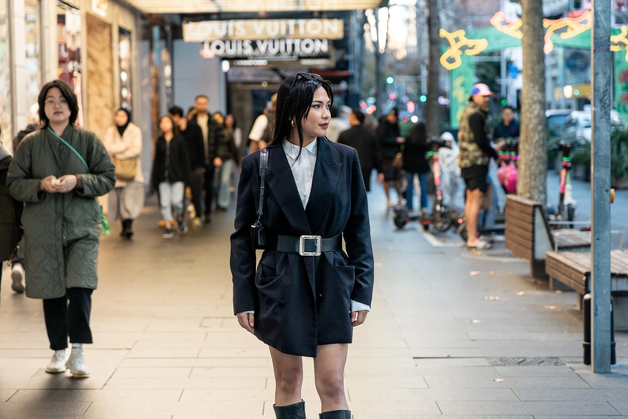 A woman with black hair wearing a black blazer dress with a white shirt underneath, black boots, and sunglasses on her head, walking in a busy outdoor shopping area with storefronts, signs, and other shoppers in the background.