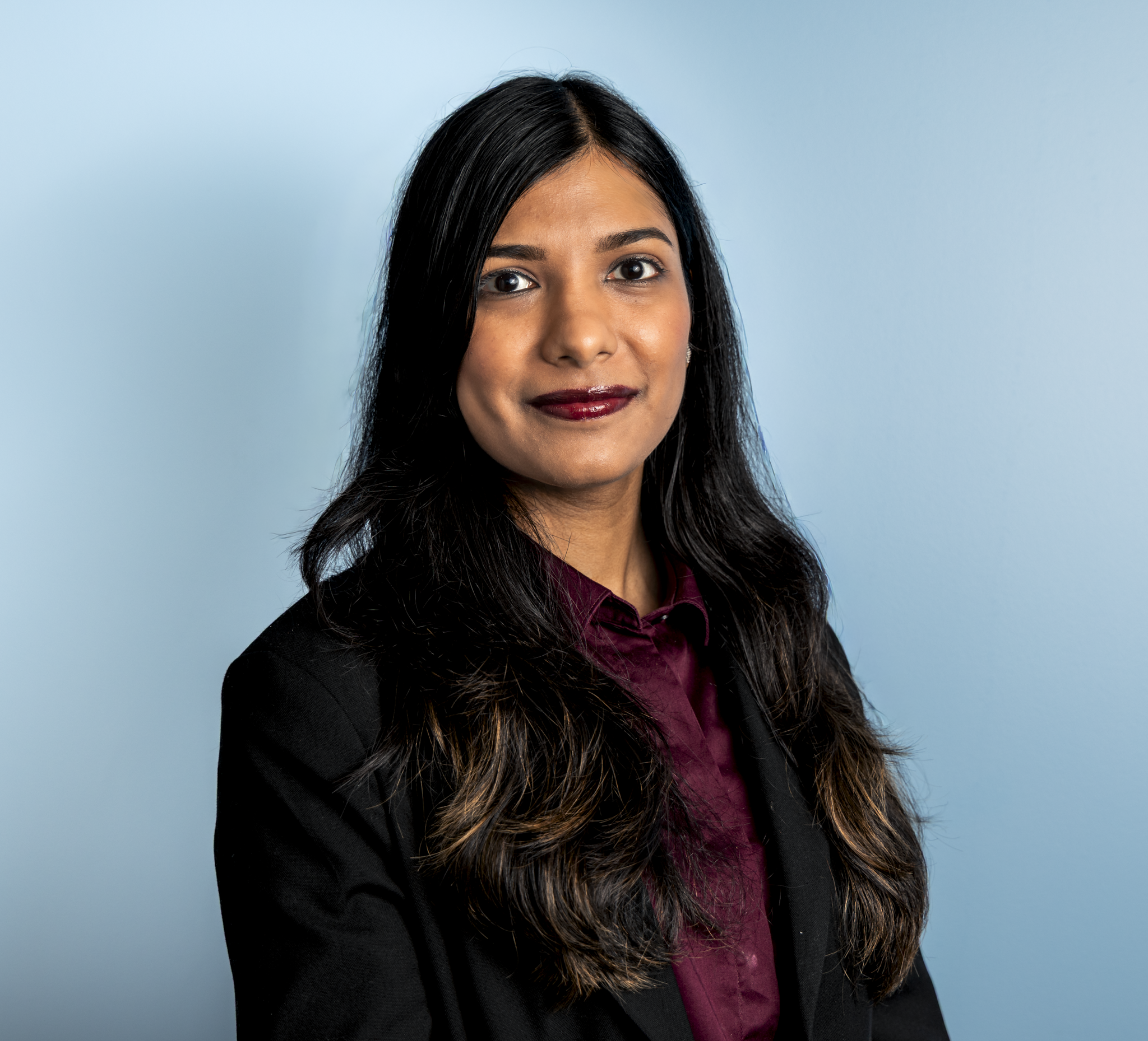 A woman with long black hair and dark lipstick wearing a black blazer and maroon shirt, standing against a light blue background.