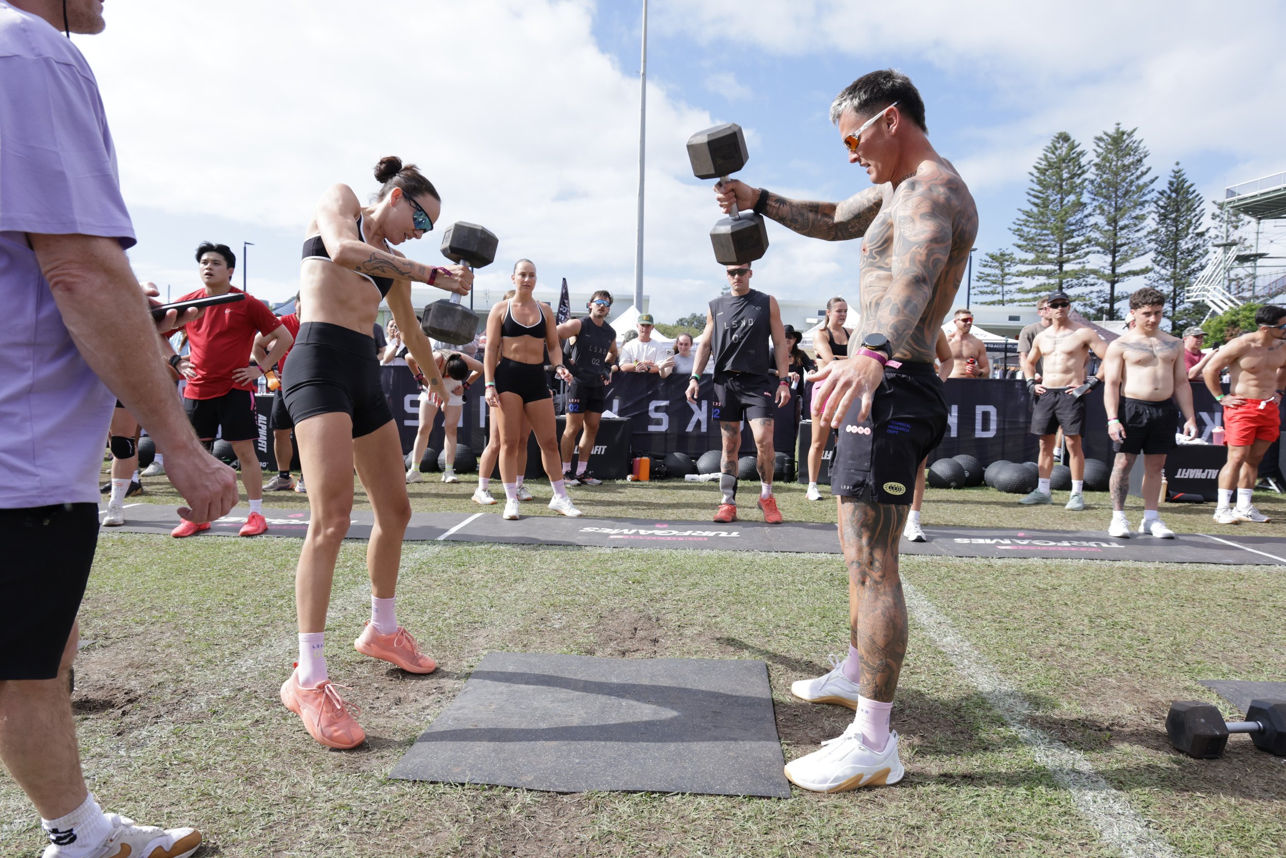 Two tattooed athletes, a man and a woman, are lifting dumbbells during a CrossFit competition outdoors on a grassy field, with other participants and spectators in the background.