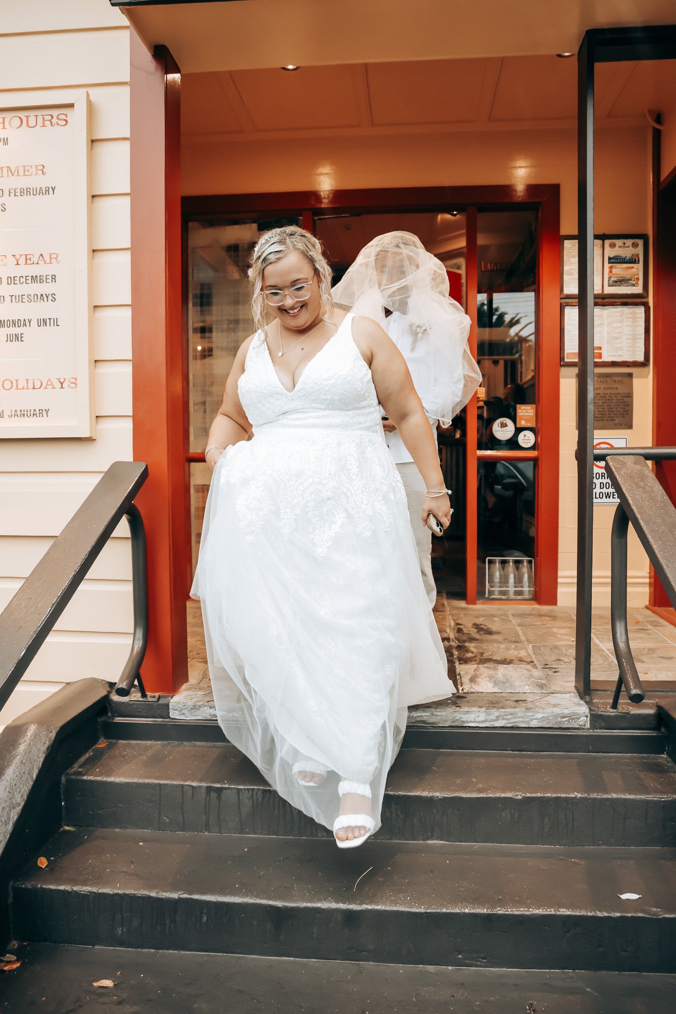 A woman in a white wedding dress with glasses and curly hair is smiling as she descends a set of black stairs outside a building with a red door, behind her is a person in a white suit with a veil.